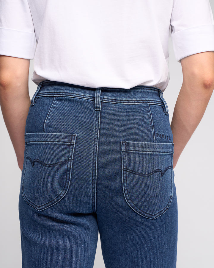 Model wears Vassalli Peace Jean - Brushed Indigo, a high-waisted wide-leg denim jean with a relaxed fit, two large back pockets, and decorative stitching, paired with a white short-sleeve shirt. Shown from the back.