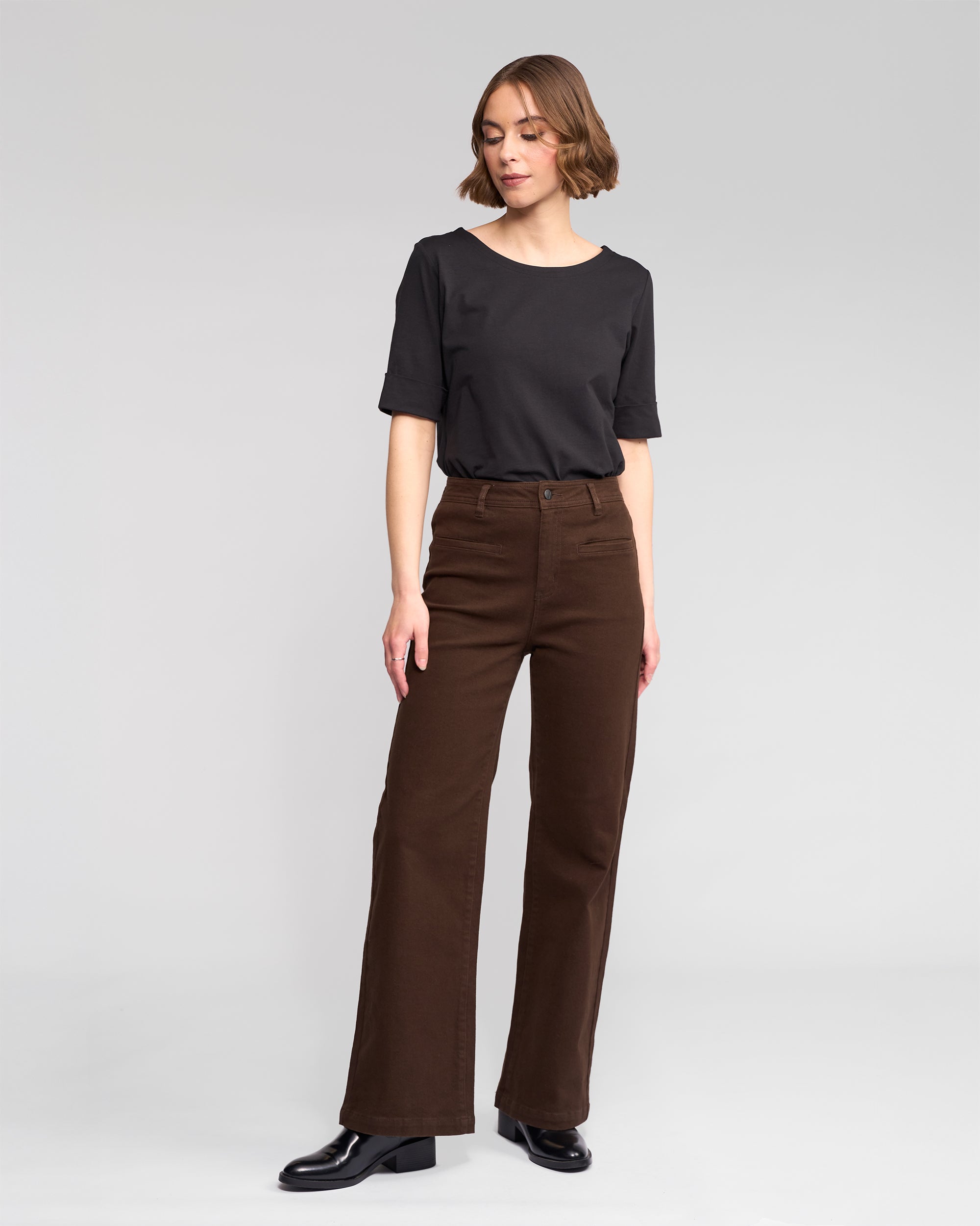 A woman stands against a plain light gray background, wearing a black short-sleeve top, black shoes, and Vassalli Peace Jean - Chocolate wide-leg denim pants. She looks slightly left with her right hand on her thigh.