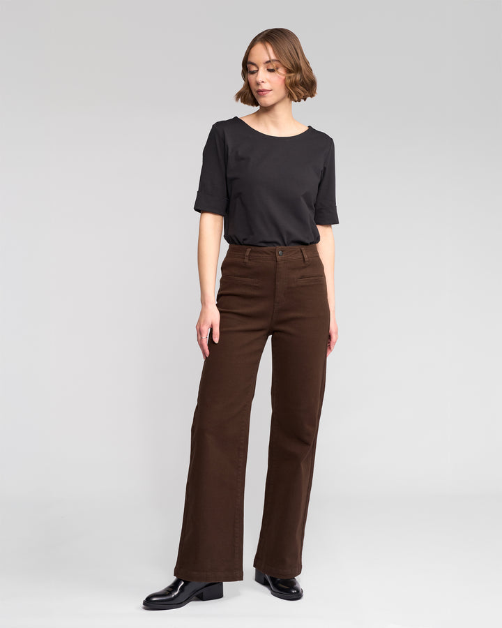 A woman stands against a plain light gray background, wearing a black short-sleeve top, black shoes, and Vassalli Peace Jean - Chocolate wide-leg denim pants. She looks slightly left with her right hand on her thigh.