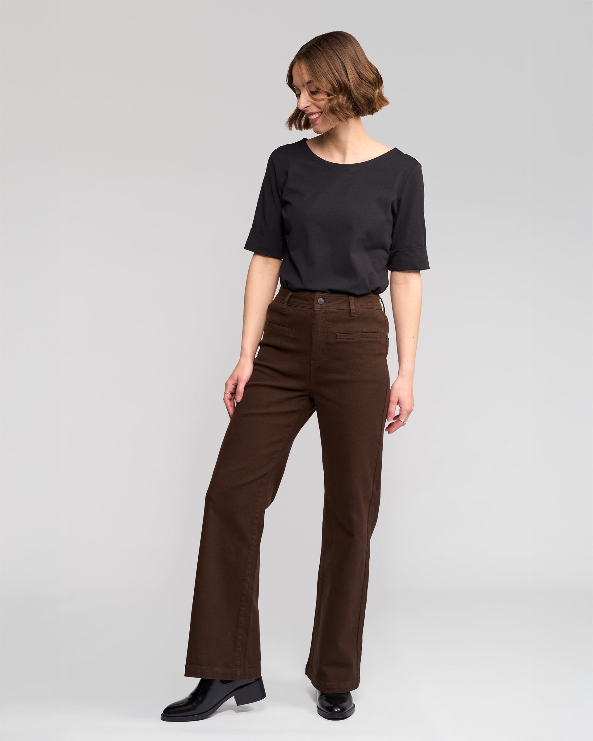 A woman with short, wavy hair stands against a light gray background, wearing the Vassalli Peace Jean in Chocolate, a black short-sleeve top, and black ankle boots. She looks down with a slight smile.