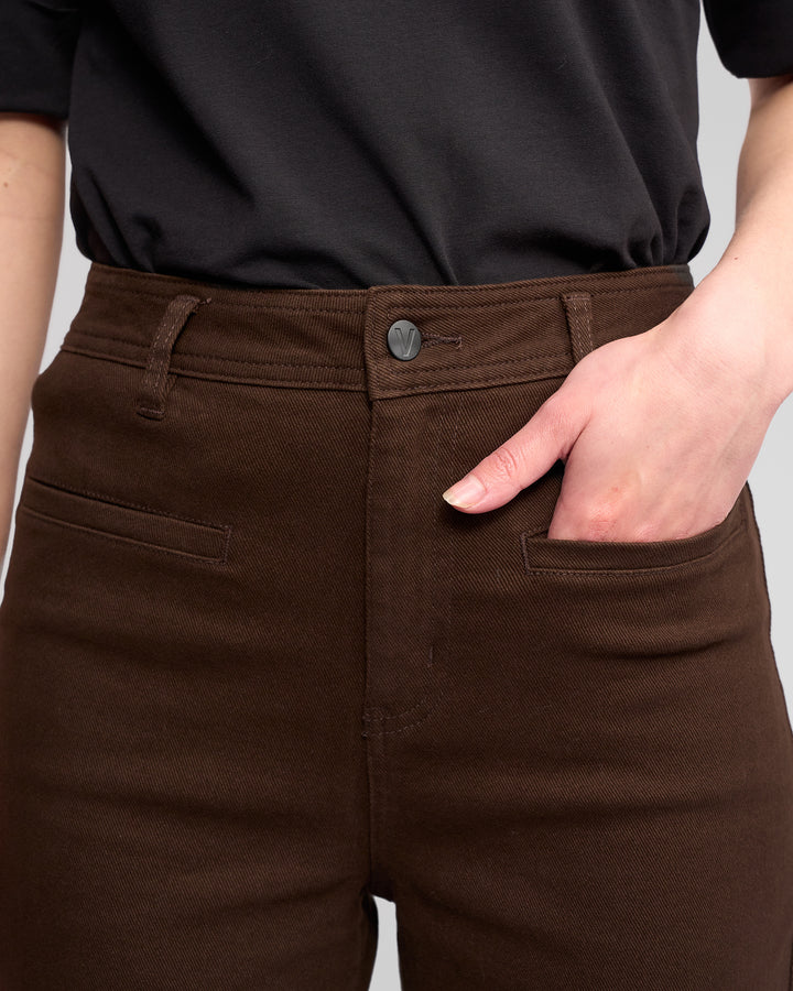 A person wearing Vassalli Peace Jean - Chocolate, styled high-waisted and wide-leg, with a black shirt has their right hand in the front pocket. The image captures the waist and part of the torso.