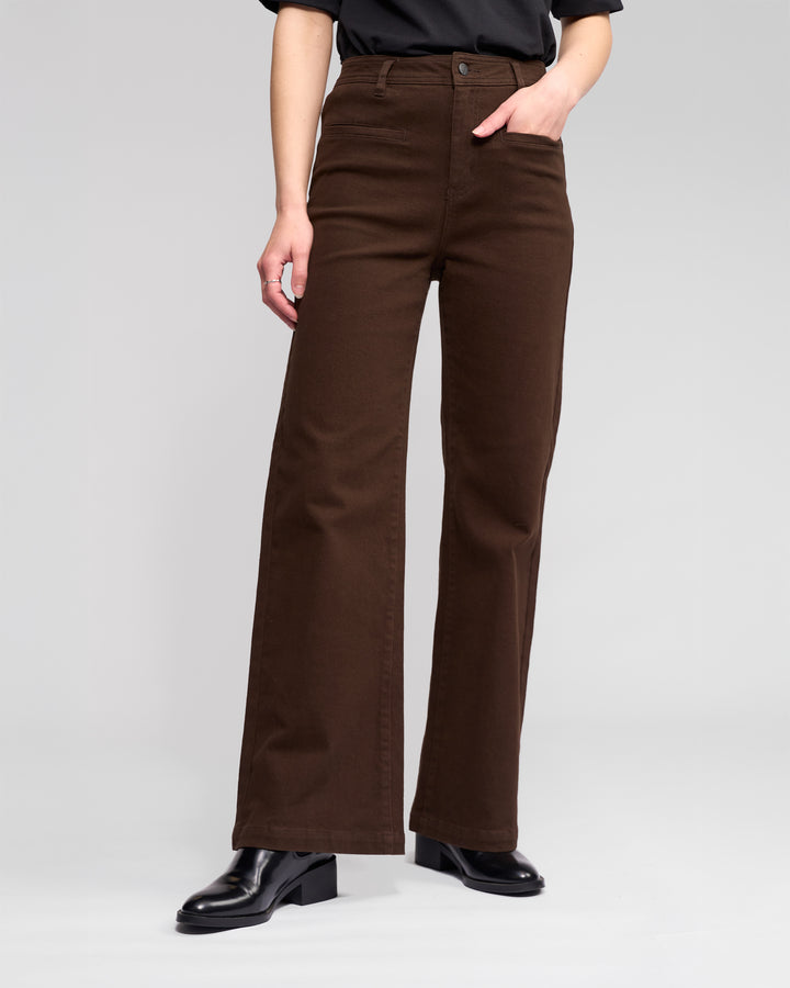 Wearing Vassalli's Peace Jean in Chocolate, paired with a black top and black ankle boots, a person stands with one hand in their pocket against a plain light gray background.