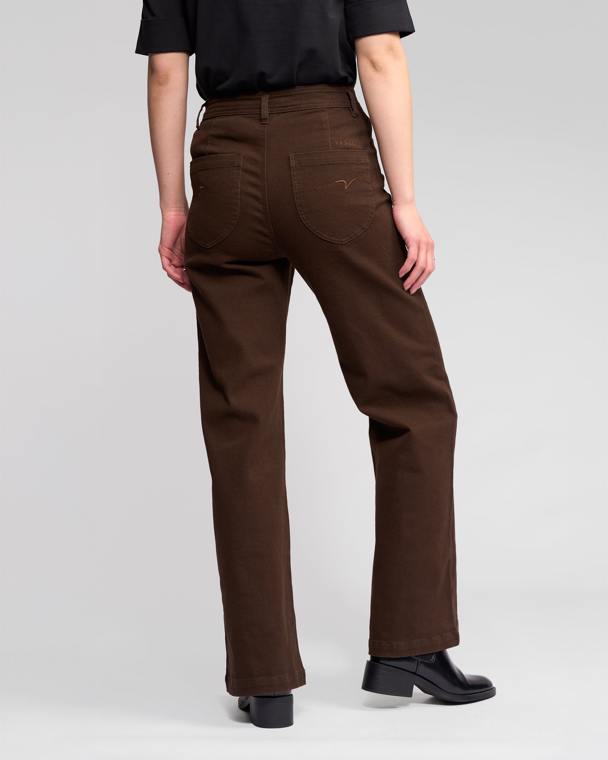A person wearing a black shirt, Vassalli's Peace Jean in Chocolate, and black shoes stands with their back to the camera against a plain light gray background.