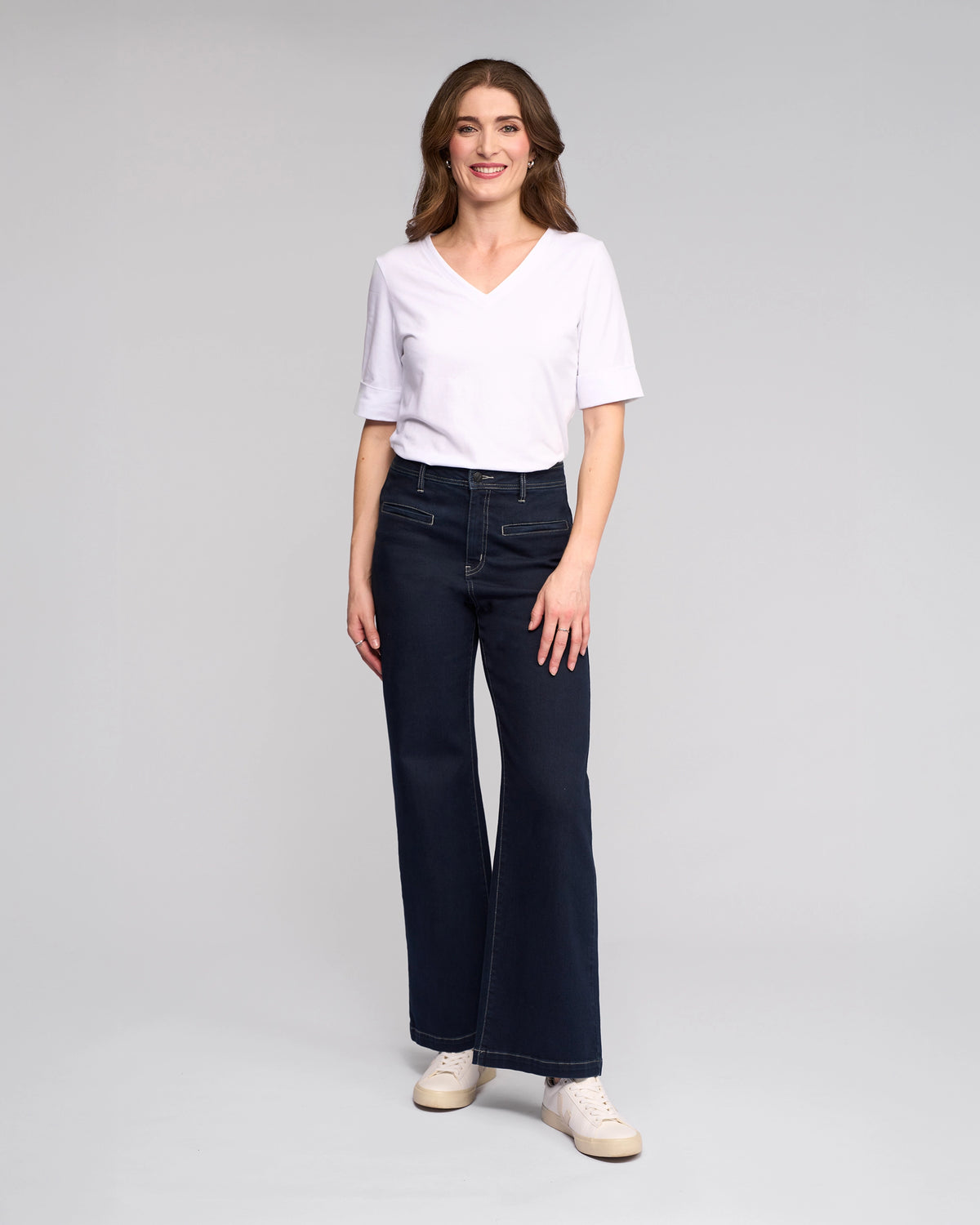 A woman with medium-length brown hair smiles against a light gray background, wearing Vassalli's Peace Jean - Ink Denim, a white V-neck tee, and white sneakers, radiating effortless style.