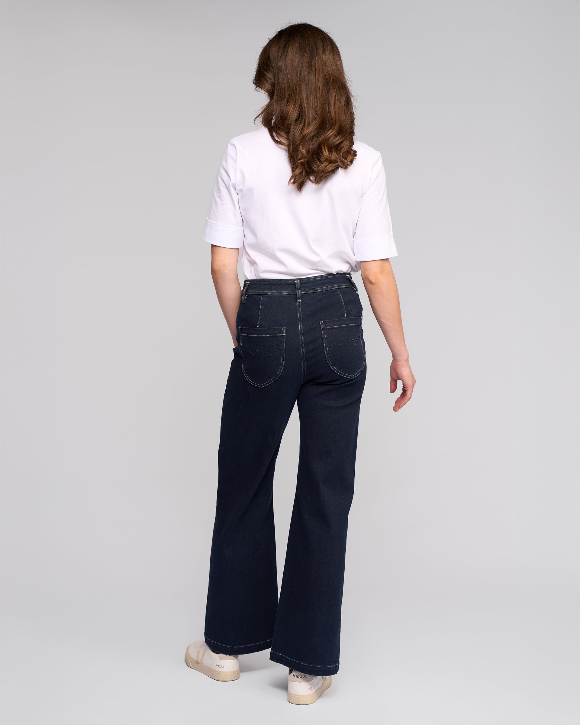 A woman with wavy brown hair, seen from behind, wears a white short-sleeve shirt and Vassalli’s Peace Jean - Ink Denim wide-leg jeans for a relaxed look, paired with white shoes against a plain light gray background.