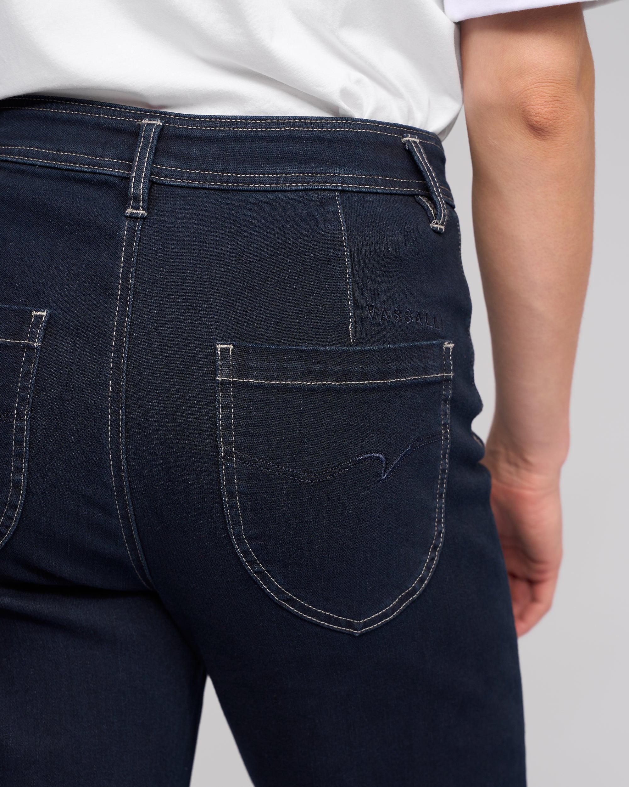 Close-up of a person wearing Vassalli Peace Jean - Ink Denim and a white shirt, highlighting the back pocket with visible stitching and the Vassalli brand name embroidered above it.