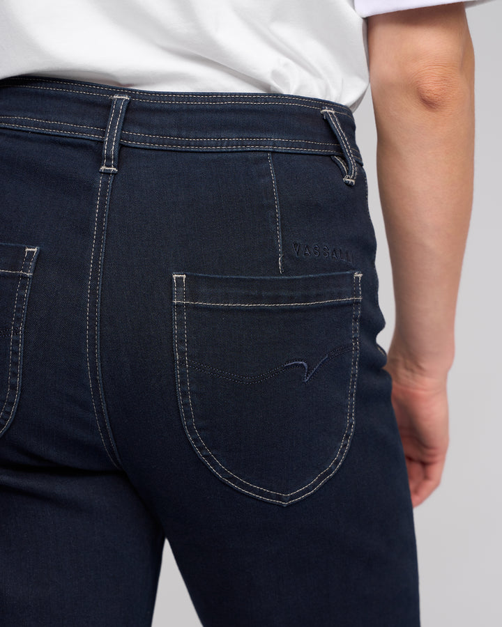Close-up of a person wearing Vassalli Peace Jean - Ink Denim and a white shirt, highlighting the back pocket with visible stitching and the Vassalli brand name embroidered above it.