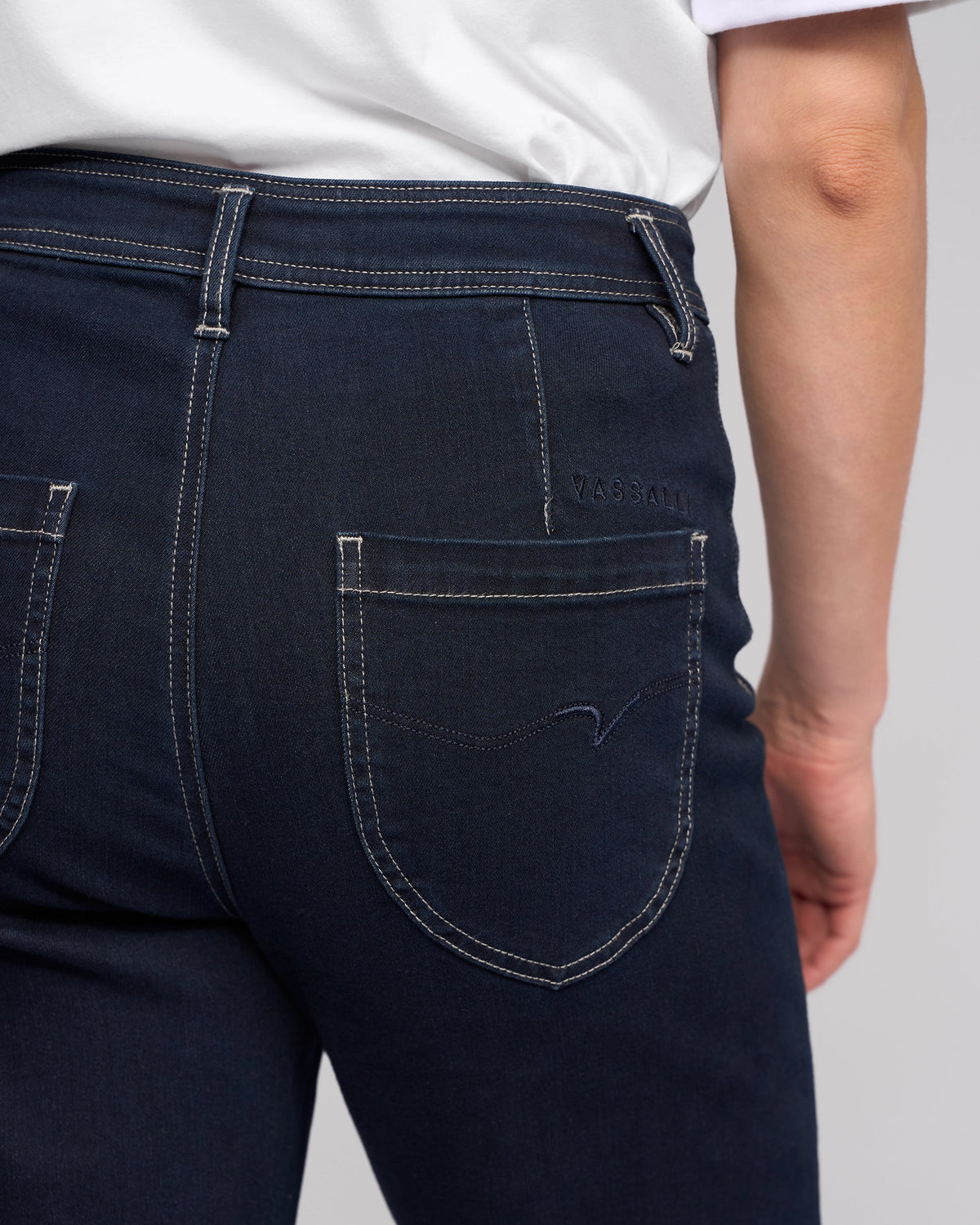 A person wearing Vassalli Peace Jean - Ink Denim and a white shirt, shown from the back waist down. The jeans feature two back pockets with visible stitching and “Vassalli” embroidered above the right pocket.