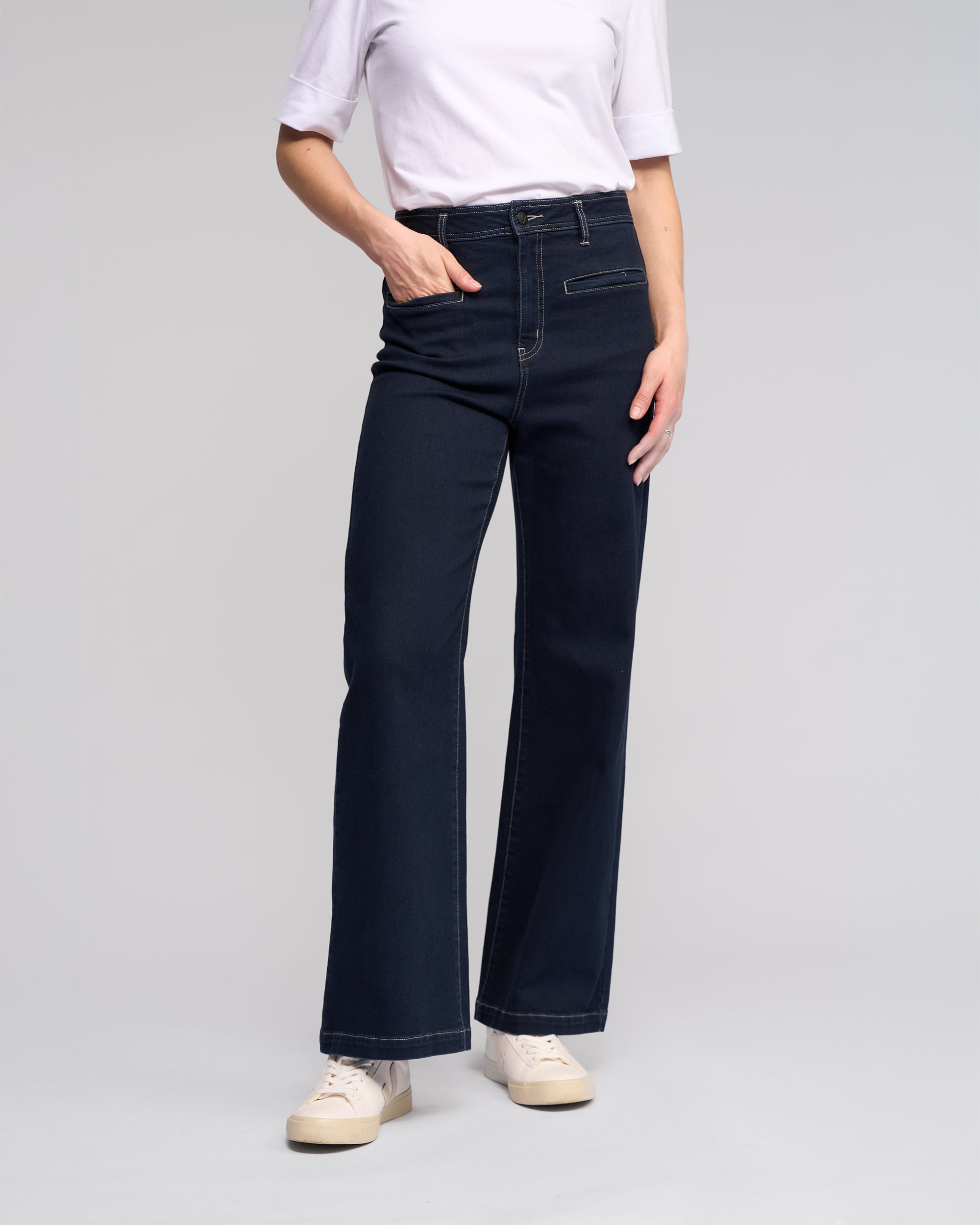 A person wearing a white short-sleeve shirt, Vassalli Peace Jean - Ink Denim with a relaxed wide-leg silhouette, and white sneakers stands against a plain light gray background, one hand in their pocket.