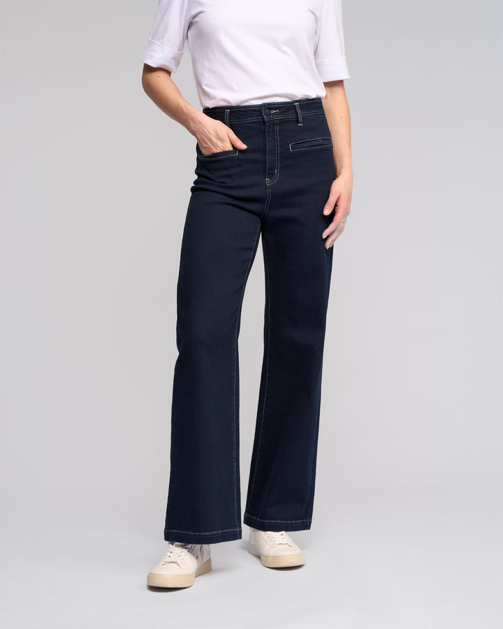 A person wearing a white short-sleeve shirt, Vassalli Peace Jean - Ink Denim with a relaxed wide-leg silhouette, and white sneakers stands against a plain light gray background, one hand in their pocket.