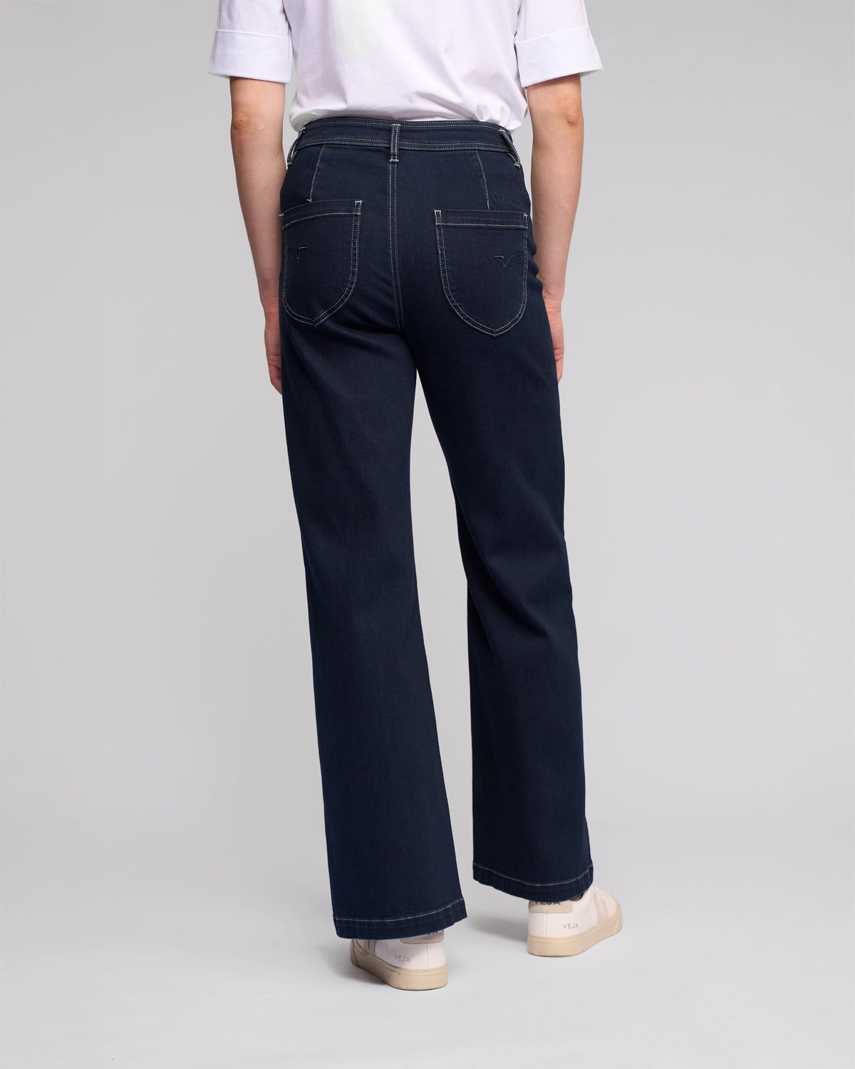 A person stands with their back to the camera, wearing a white t-shirt, Vassalli Peace Jean - Ink Denim high-waisted women's wide leg jeans, and light-colored shoes, set against a plain light background.