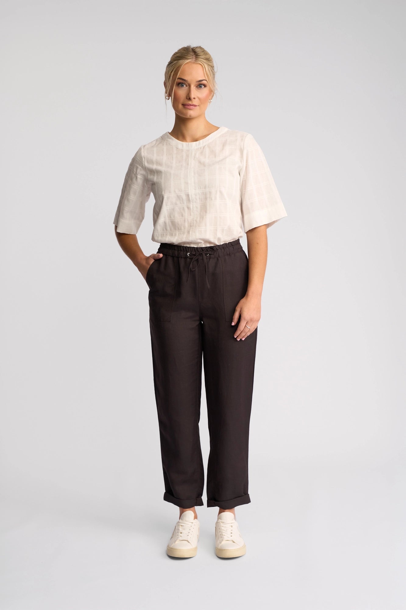 A person stands against a plain light background, wearing a white short-sleeve top, Vassalli’s Oxford Pant in Chocolate crafted from a linen-viscose blend, and white sneakers, with one hand in their pocket and a neutral expression.