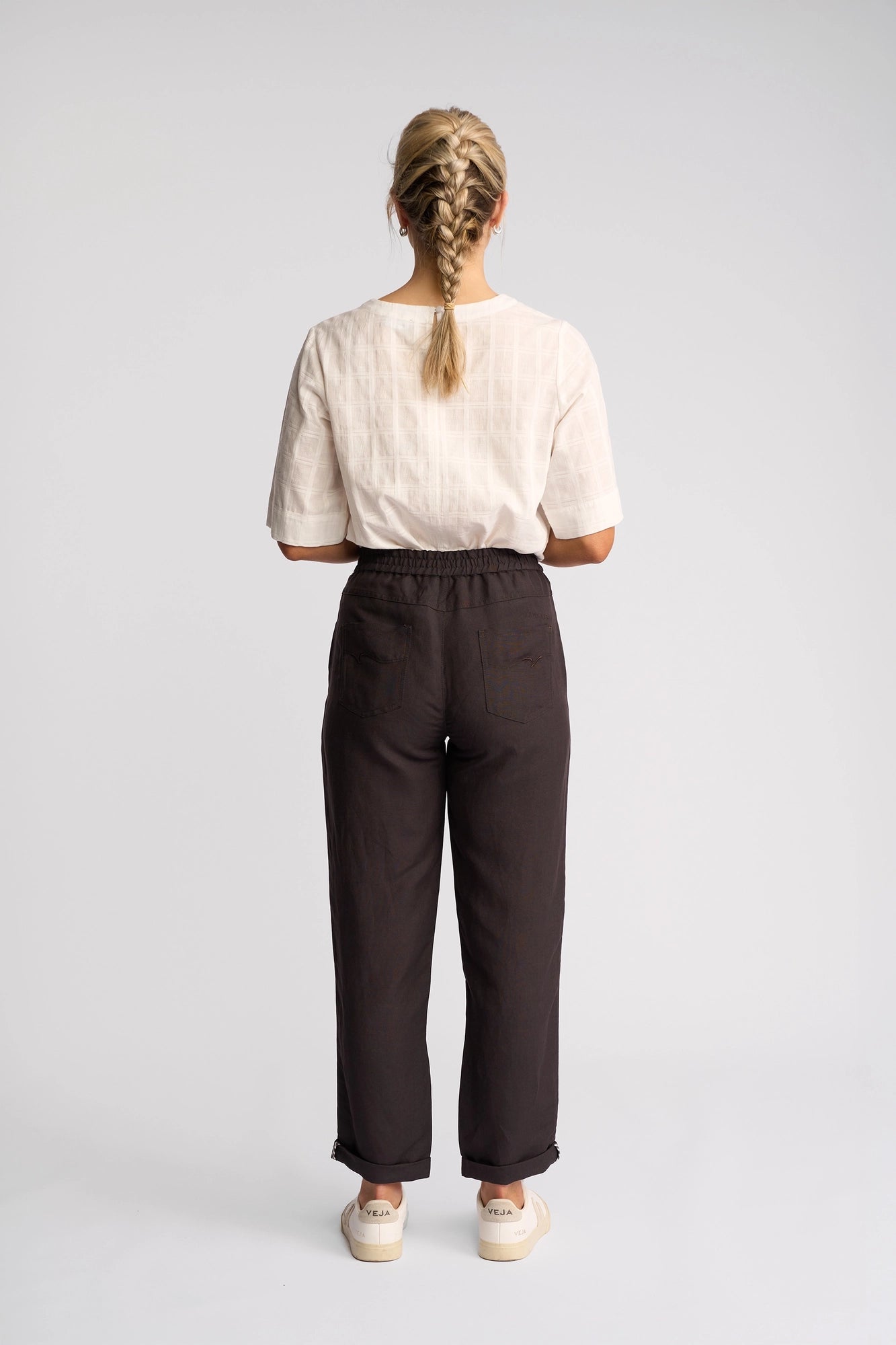 A person with blonde hair in two braids stands facing away, wearing a light short-sleeve top, the Vassalli Oxford Pant in Chocolate crafted from a linen-viscose blend, and white shoes, against a plain white background.