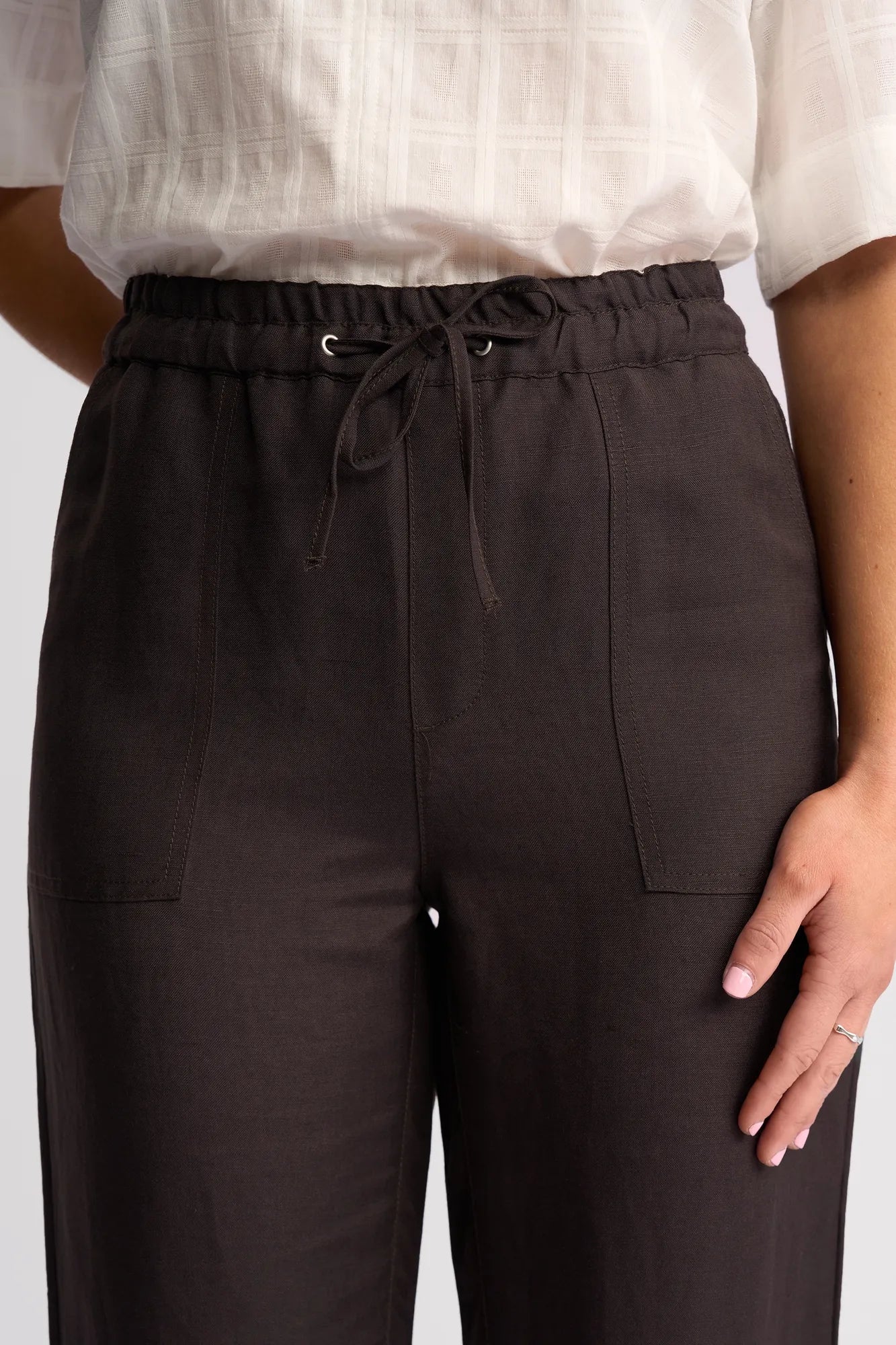A person wears a white textured short-sleeve shirt paired with the Vassalli Oxford Pant in Chocolate, a high-waisted dark brown casual summer pant featuring front patch pockets. Only the torso and part of one arm are visible.