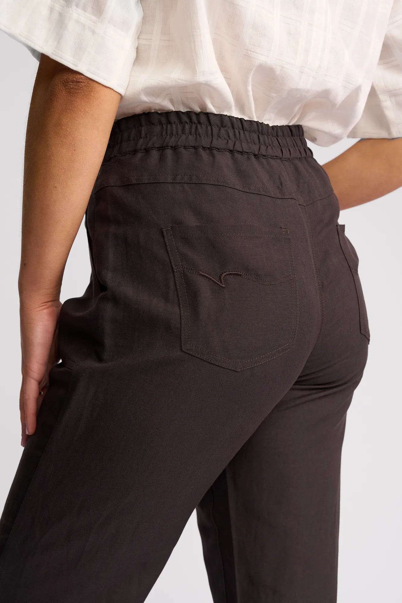Here’s a rewritten version using the provided product data: A close-up of a person wearing Vassalli’s Oxford Pant - Chocolate, featuring back pockets and paired with a white checkered short-sleeve shirt; the image highlights the elastic waistband, seam details, and the soft linen-viscose blend that makes these casual summer pants both comfortable and stylish.