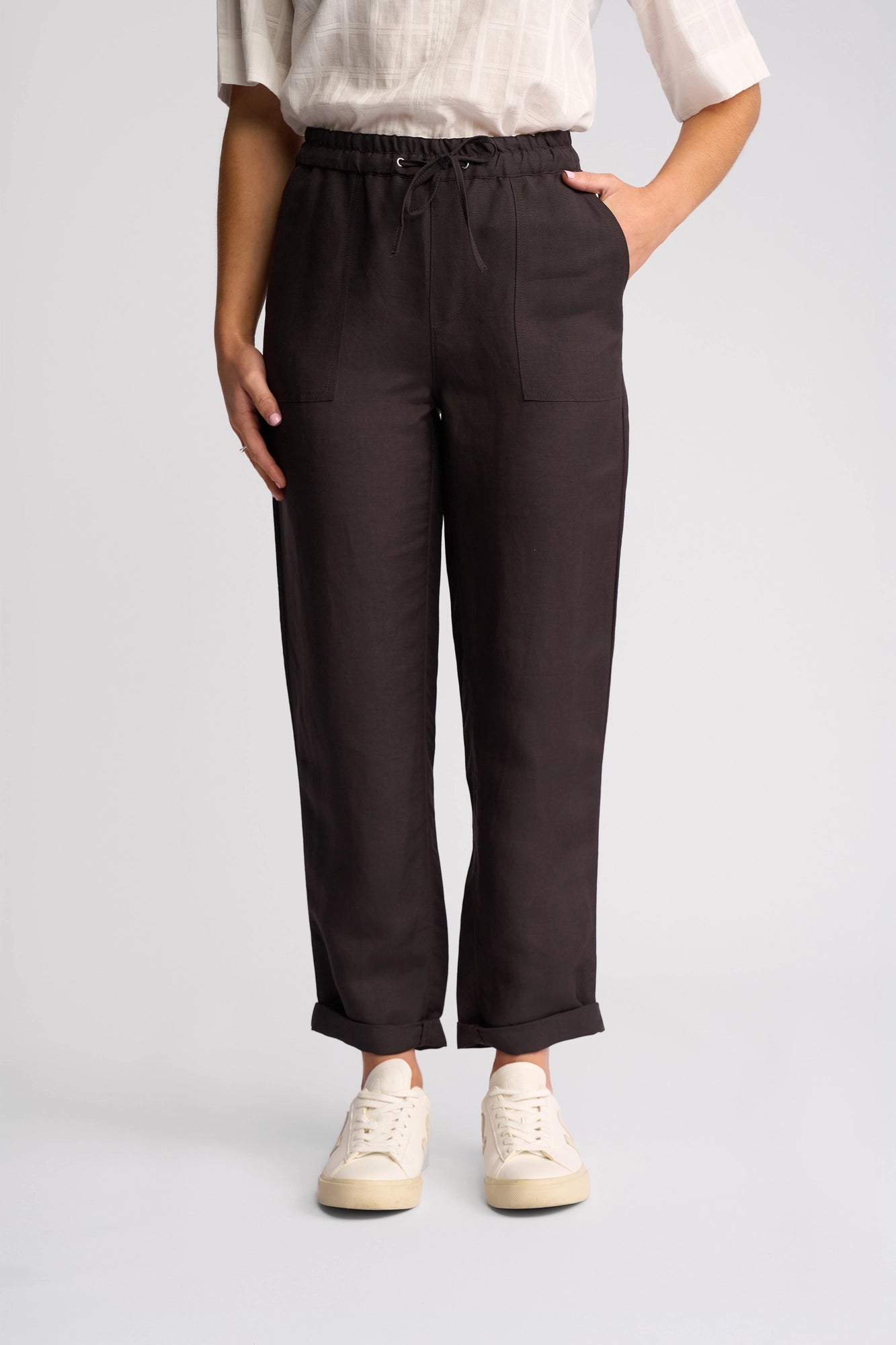 A person is wearing Vassalli’s Oxford Pant in Chocolate—a pair of black cuffed pants with front pockets—styled with a white short-sleeve shirt and crisp white sneakers. Made from a soft linen-viscose blend, these casual summer pants feature a relaxed fit, and the model stands with one hand in their pocket against a plain light gray background.