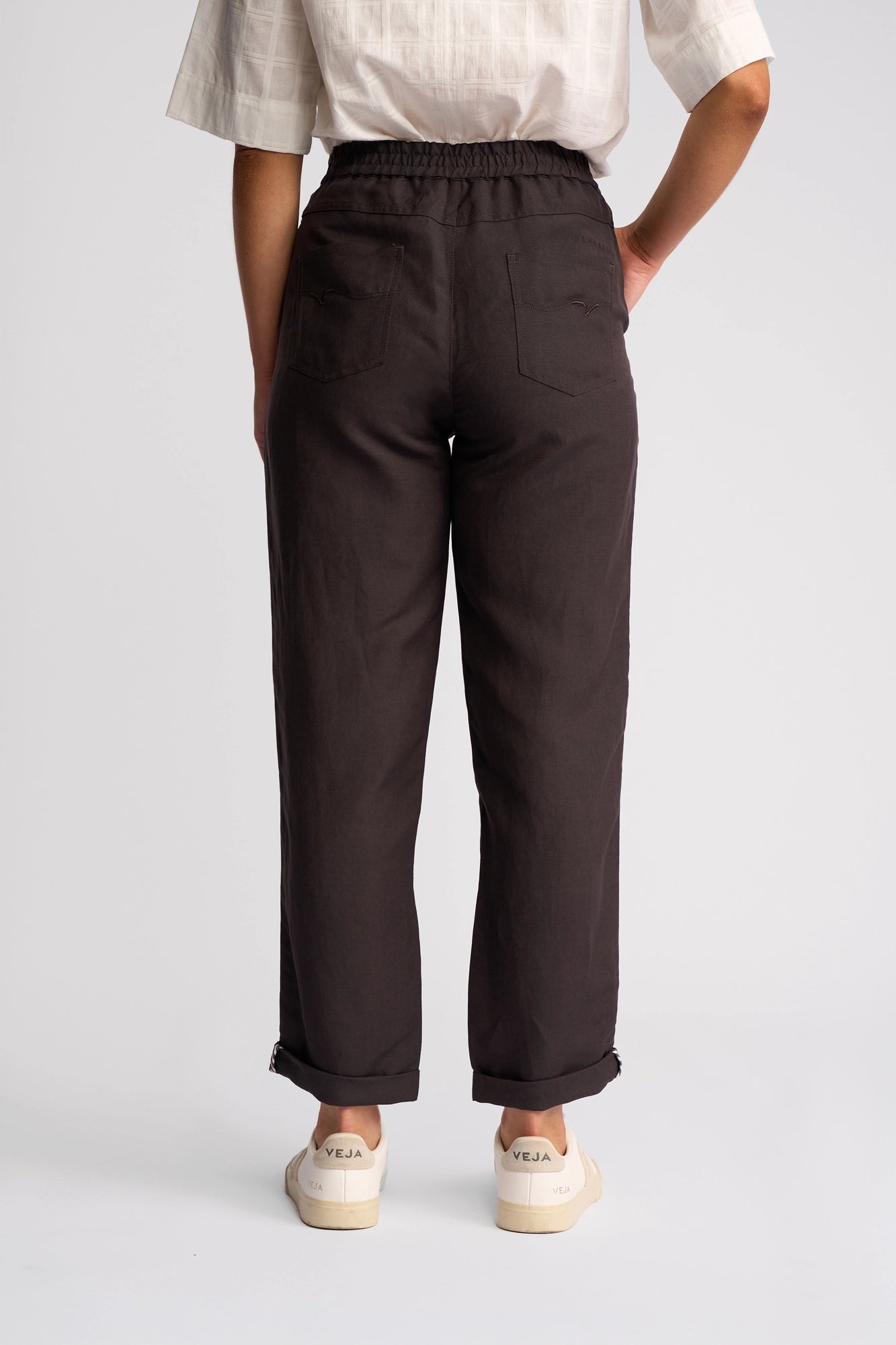 A person stands facing away against a plain, light background, wearing a white short-sleeve shirt, Vassalli’s Oxford Pant in Chocolate—a dark high-waisted casual summer pant made from a linen-viscose blend with back pockets—slightly cuffed at the ankles, and white Veja sneakers.