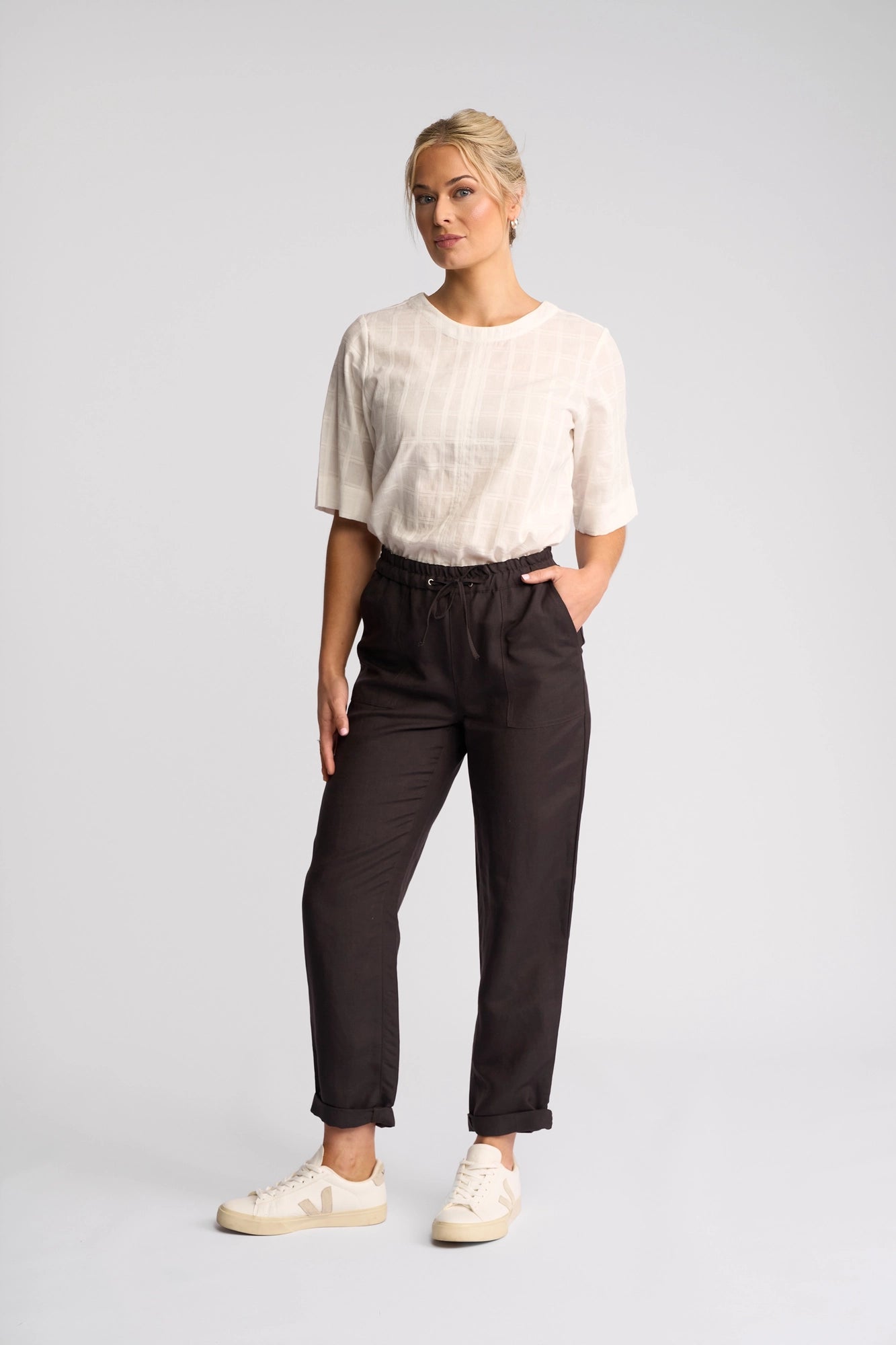 A woman stands against a plain background, wearing a white short-sleeve top, Vassalli’s Oxford Pant in Chocolate—crafted from a linen-viscose blend—and white sneakers, with one hand in her pocket and the other hanging by her side.