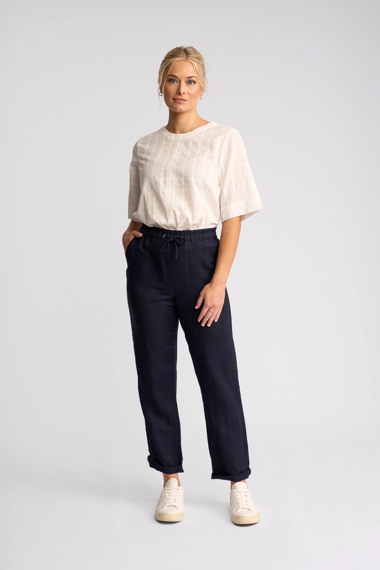 A person stands against a plain light background, wearing a light short-sleeve top, Vassalli Oxford Pant - Ink in a casual summer style made from a comfortable linen-viscose blend, and white sneakers. Their hands are in their pockets and they are looking forward with a neutral expression.
