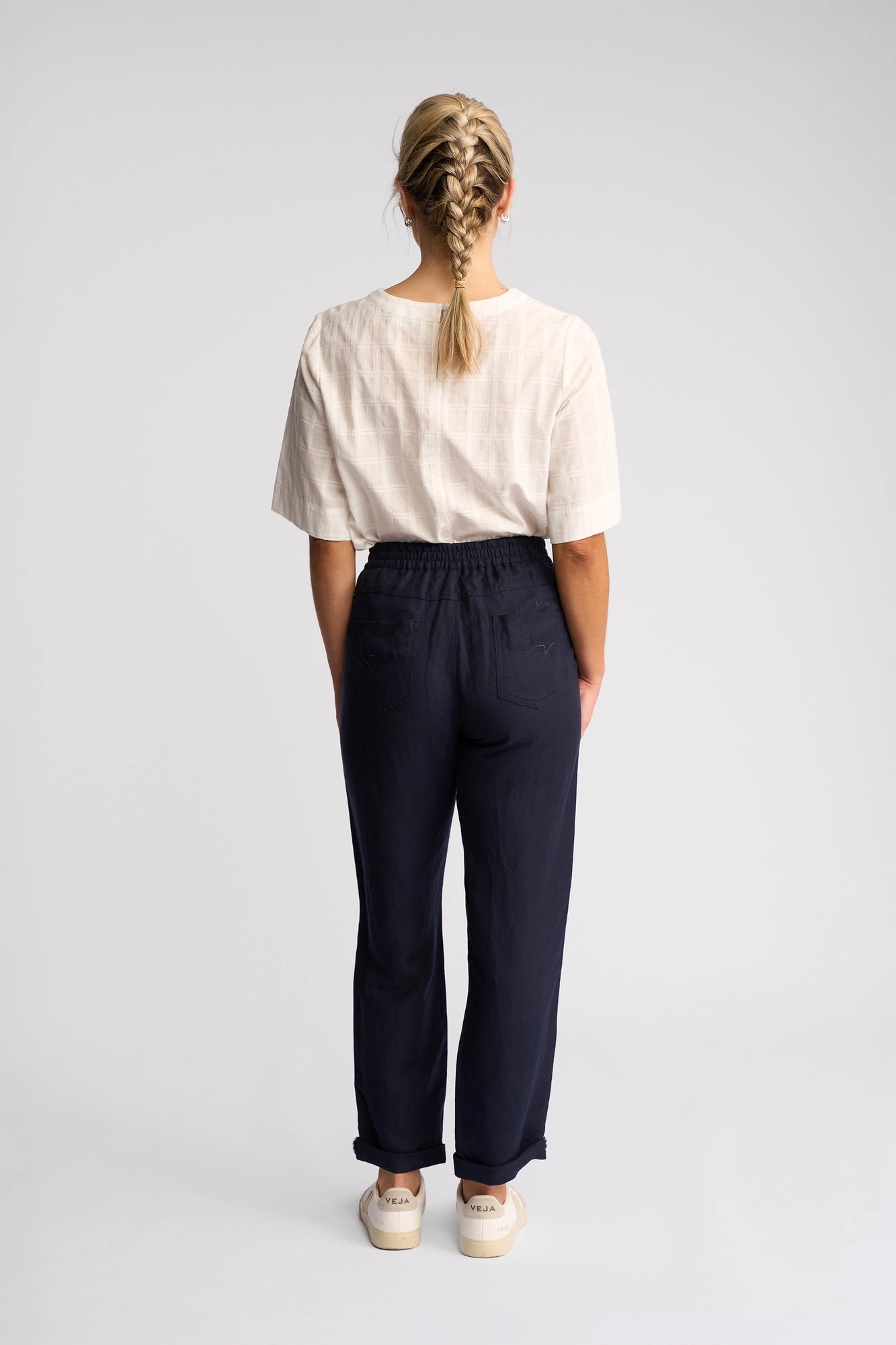 A person with blond hair in a braid stands facing away, wearing a short-sleeve beige top, Vassalli Oxford Pant - Ink, and light-colored shoes against a plain white background.