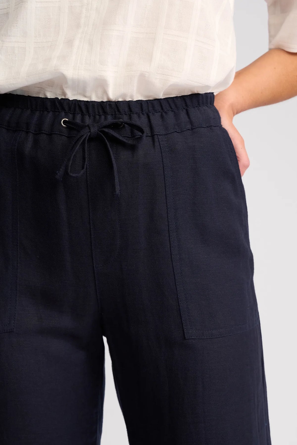 A person wearing a white textured top and Vassalli Oxford Pant - Ink, crafted from a comfortable linen-viscose blend, with hands in pockets, shown from waist to mid-thigh.