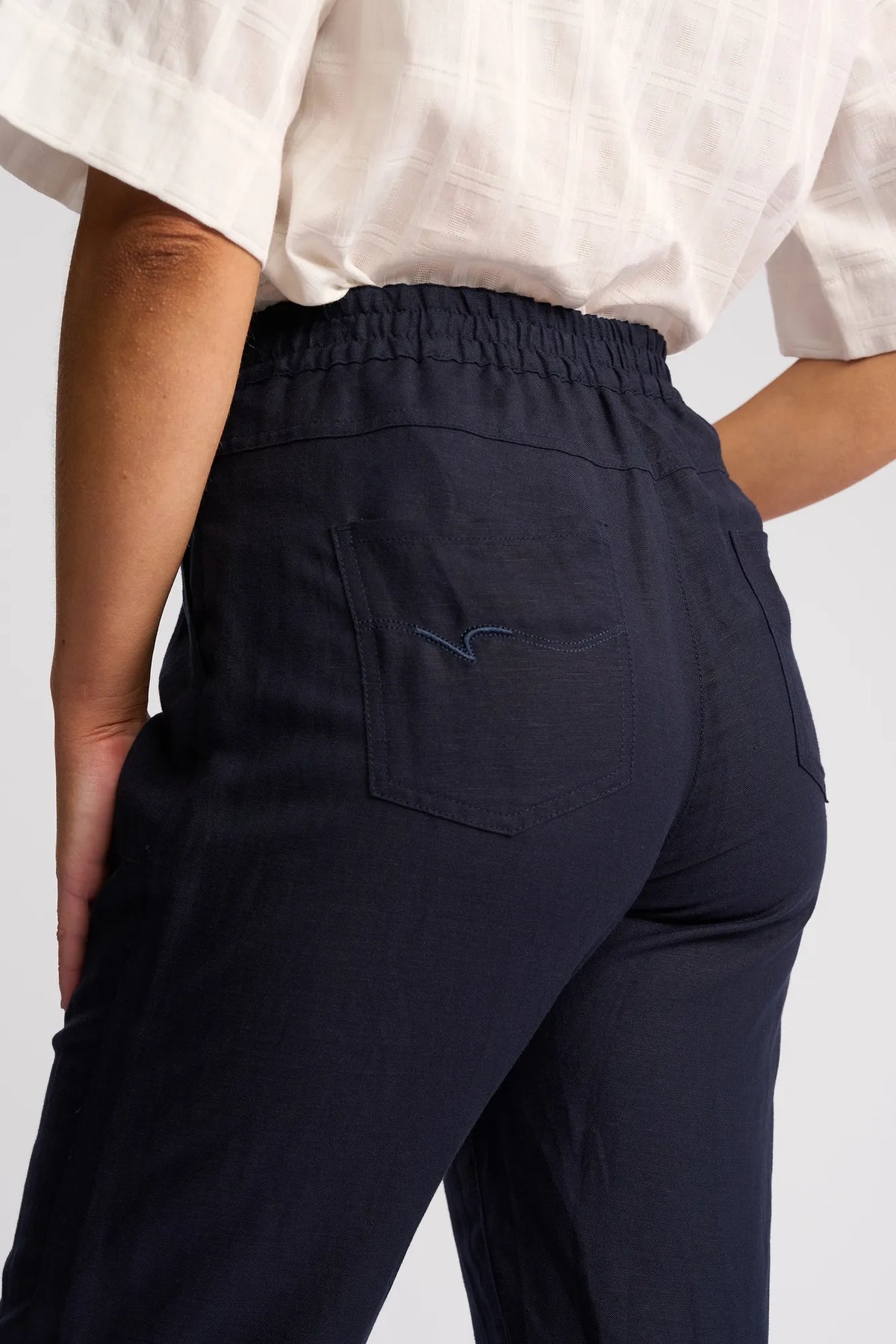 A person wearing the Vassalli Oxford Pant - Ink, a high-waisted, loose-fitting black pant with a back pocket and elastic waistband, shown from the waist down. Crafted from a breathable linen-viscose blend, these casual summer pants offer effortless style and comfort.