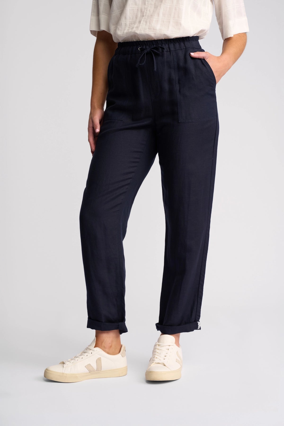 A person wearing the Vassalli Oxford Pant - Ink, crafted in a comfortable linen-viscose blend, pairs it with a white patterned shirt and cream sneakers. They stand with one hand in their pocket against a plain light background—an ideal choice for effortless summer pant style.