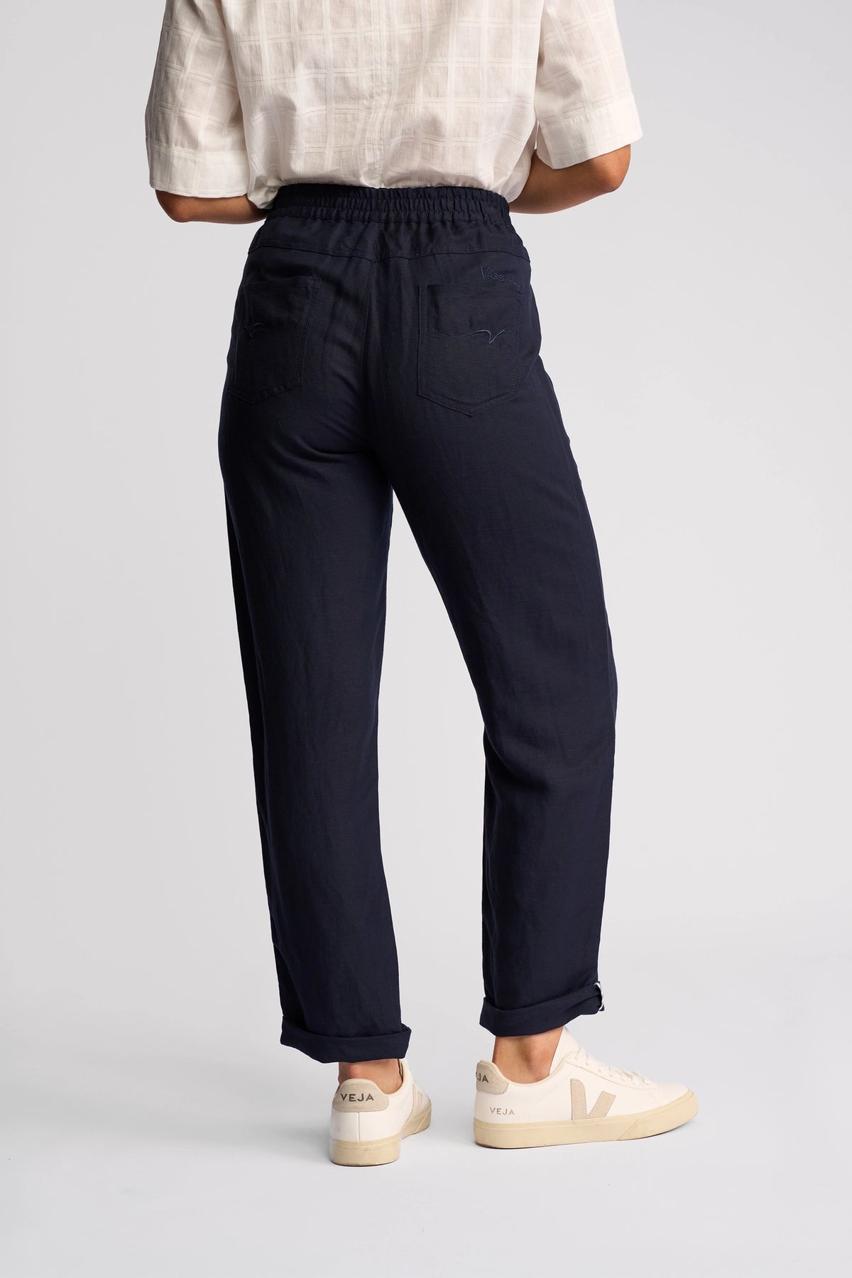 A person stands with their back to the camera, wearing a white short-sleeve shirt, beige Veja sneakers, and Vassalli’s Oxford Pant in Ink—a dark navy style crafted from a linen-viscose blend with back pockets. The background is plain and light-colored.