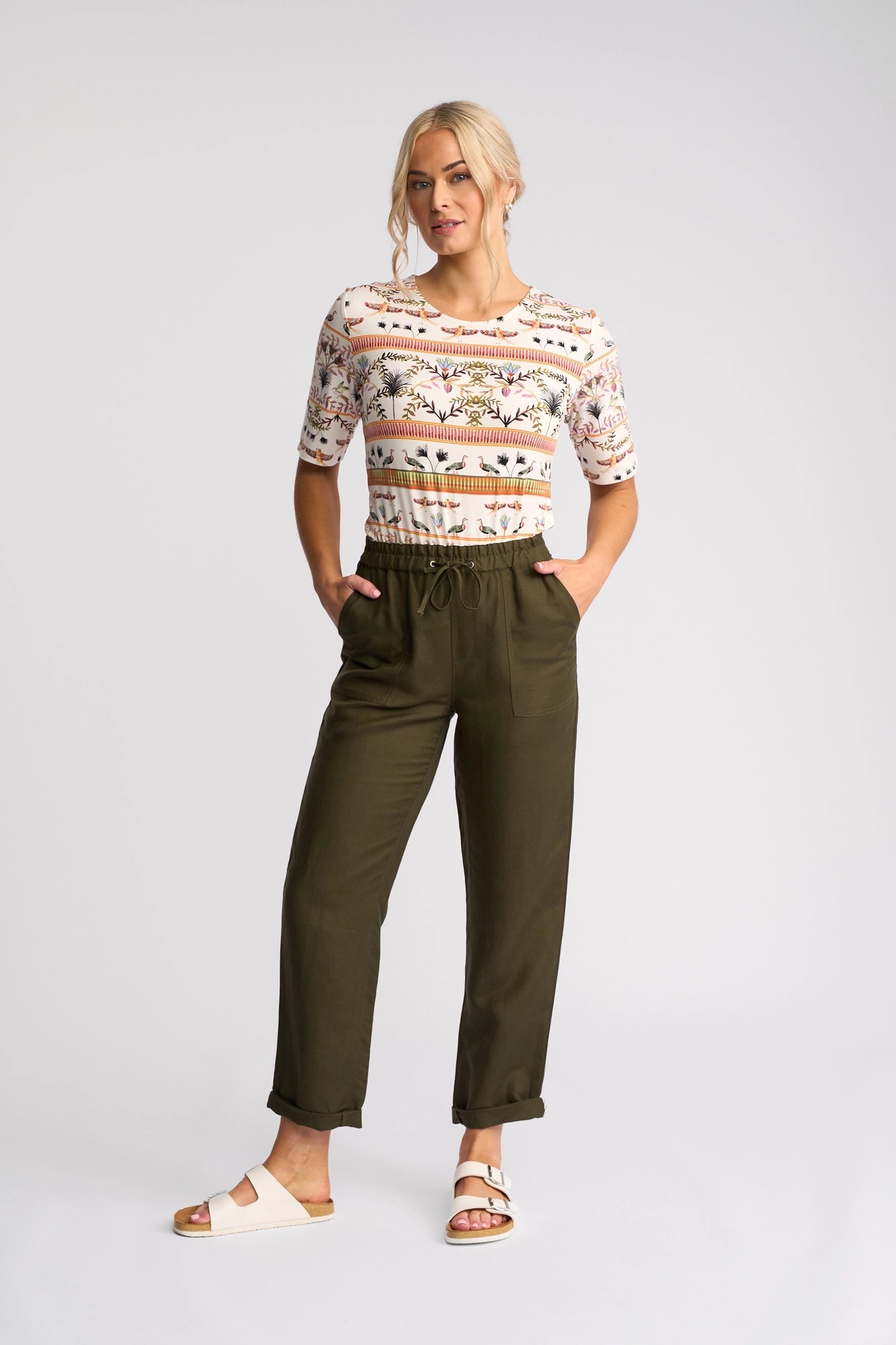 A woman poses against a plain background, hands in her pockets, wearing the Vassalli Oxford Pant in Khaki—a comfortable linen-viscose blend—paired with a patterned short-sleeve top and white sandals. Her blonde hair is styled in a loose updo for an effortlessly casual look.
