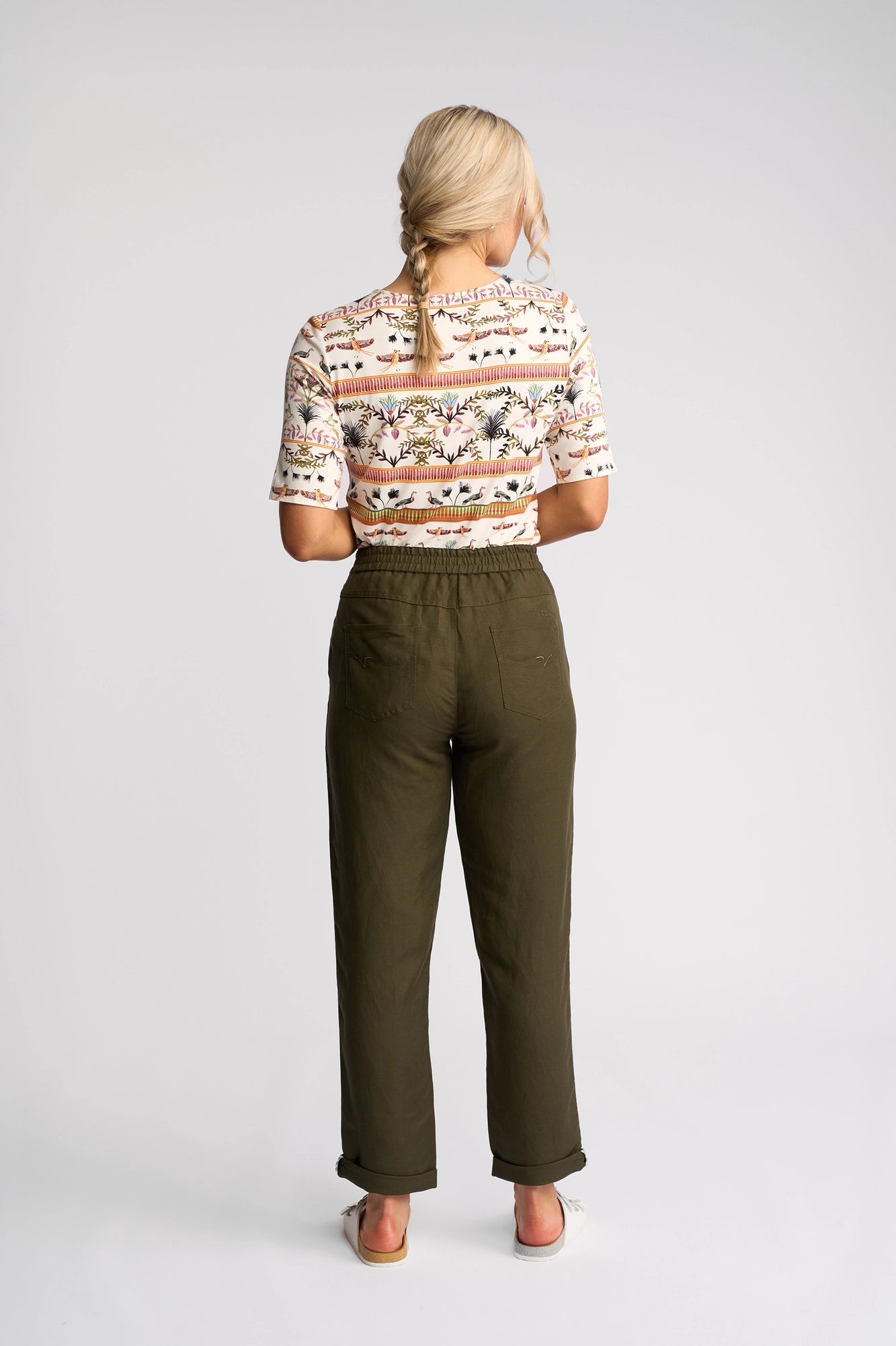 A woman with blonde braided hair stands facing away, wearing a patterned short-sleeve shirt, white shoes, and the Vassalli Oxford Pant in Khaki—crafted from a linen-viscose blend—against a plain white background.