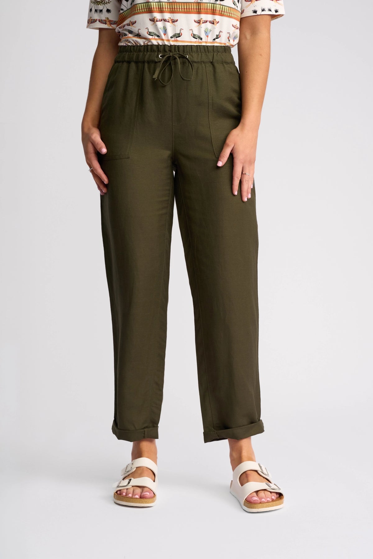 A person wearing Vassalli’s Oxford Pant in khaki, paired with a patterned shirt and white sandals, stands against a plain light background. Only the lower half of the body is visible.
