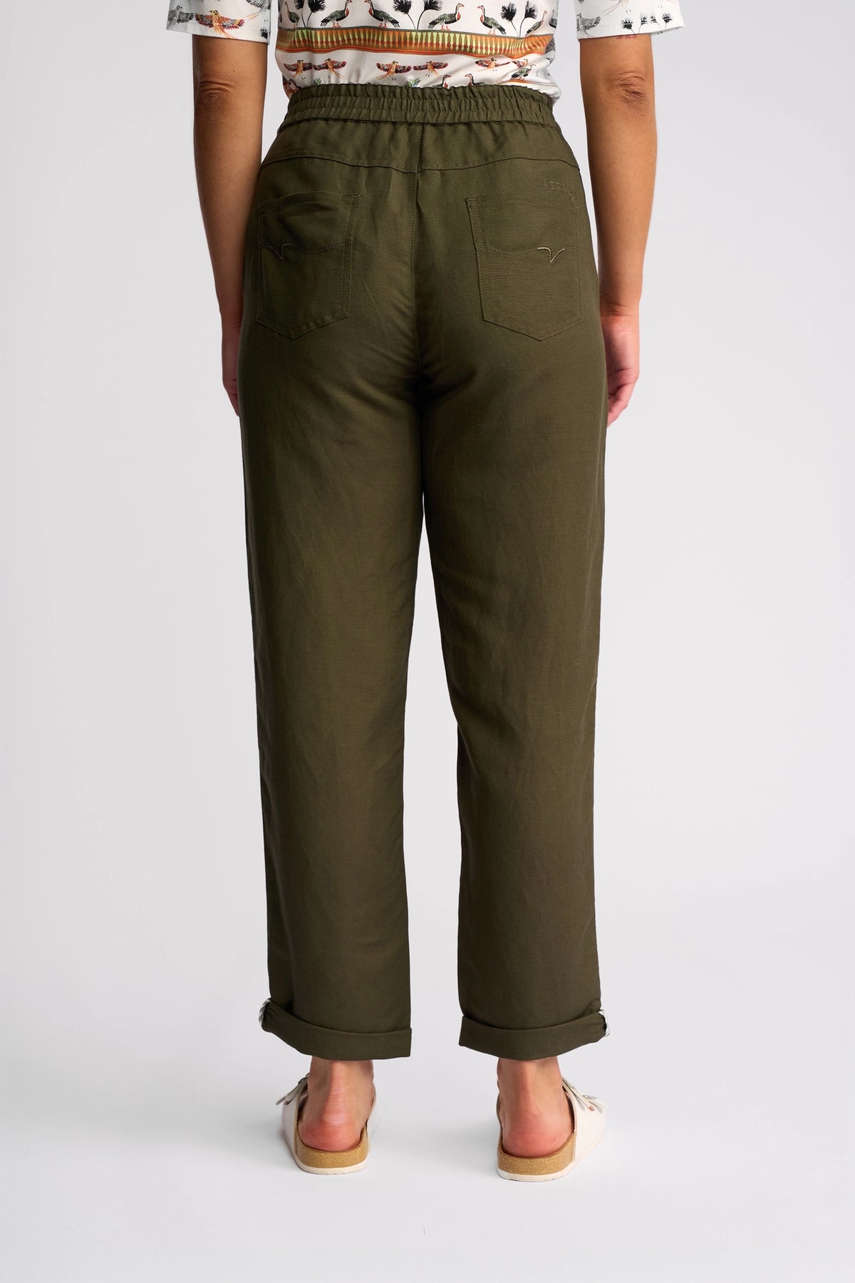 A person wearing Vassalli’s Oxford Pant in Khaki, crafted from a linen-viscose blend, with cuffed hems and two back pockets, stands facing away alongside a patterned shirt. The plain white background emphasizes the back view of the pants.
