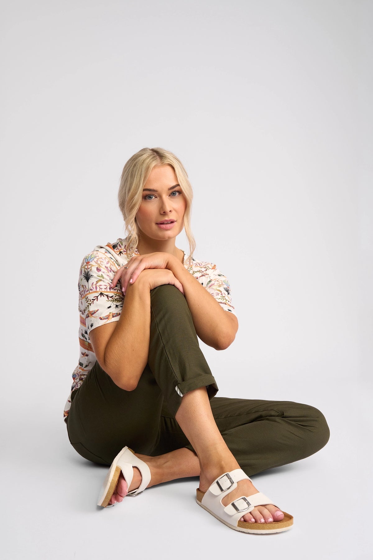 A woman with blonde hair, dressed in a patterned shirt, Vassalli Oxford Pant - Khaki, and white sandals, sits on the floor with one knee up and her hand resting on her chin against a plain background.