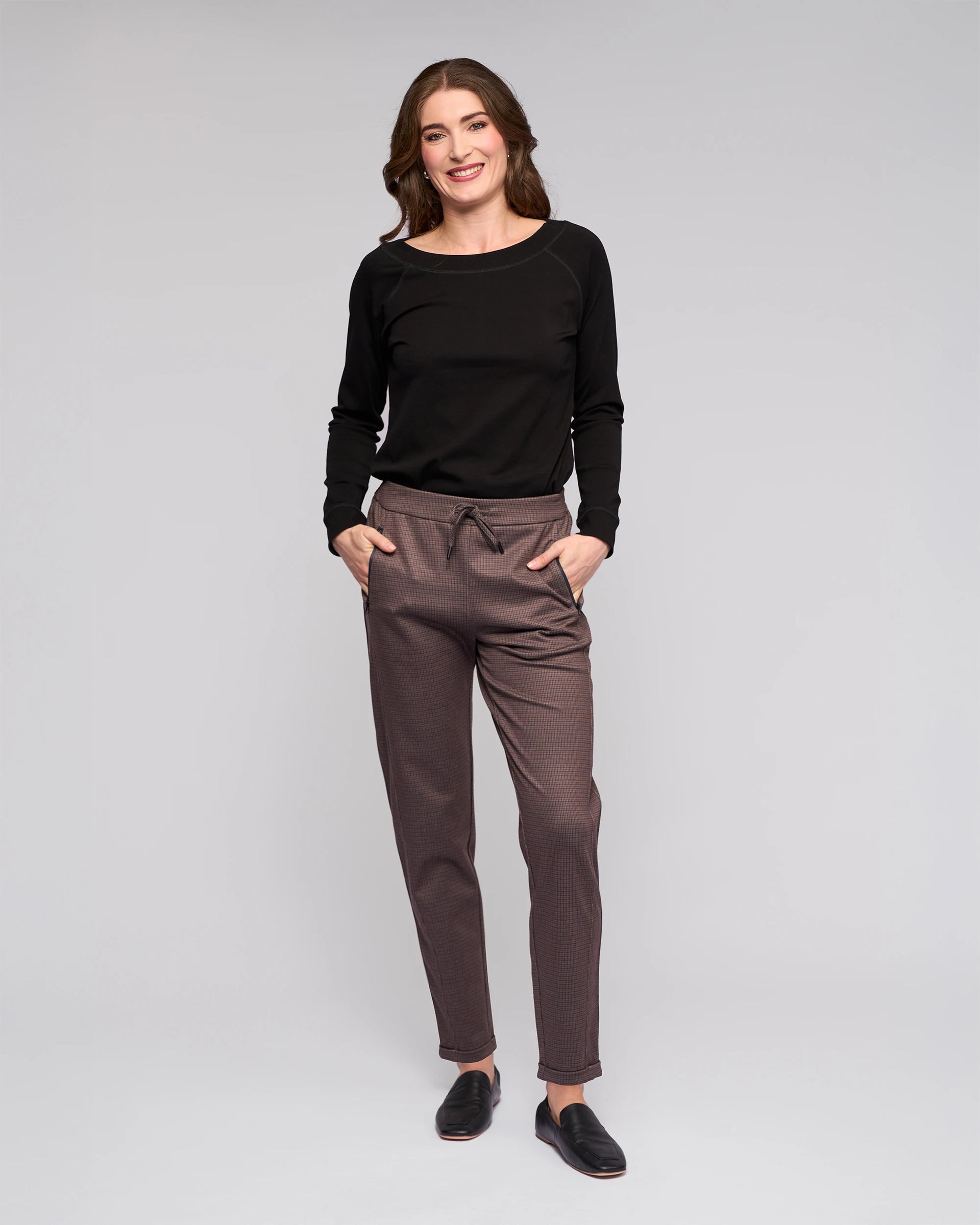 A woman with wavy brown hair smiles with her hands in her pockets, wearing Vassalli's Brooklyn Pant in Chocolate and a black long-sleeve top, paired with black slip-on shoes against a plain light gray background.