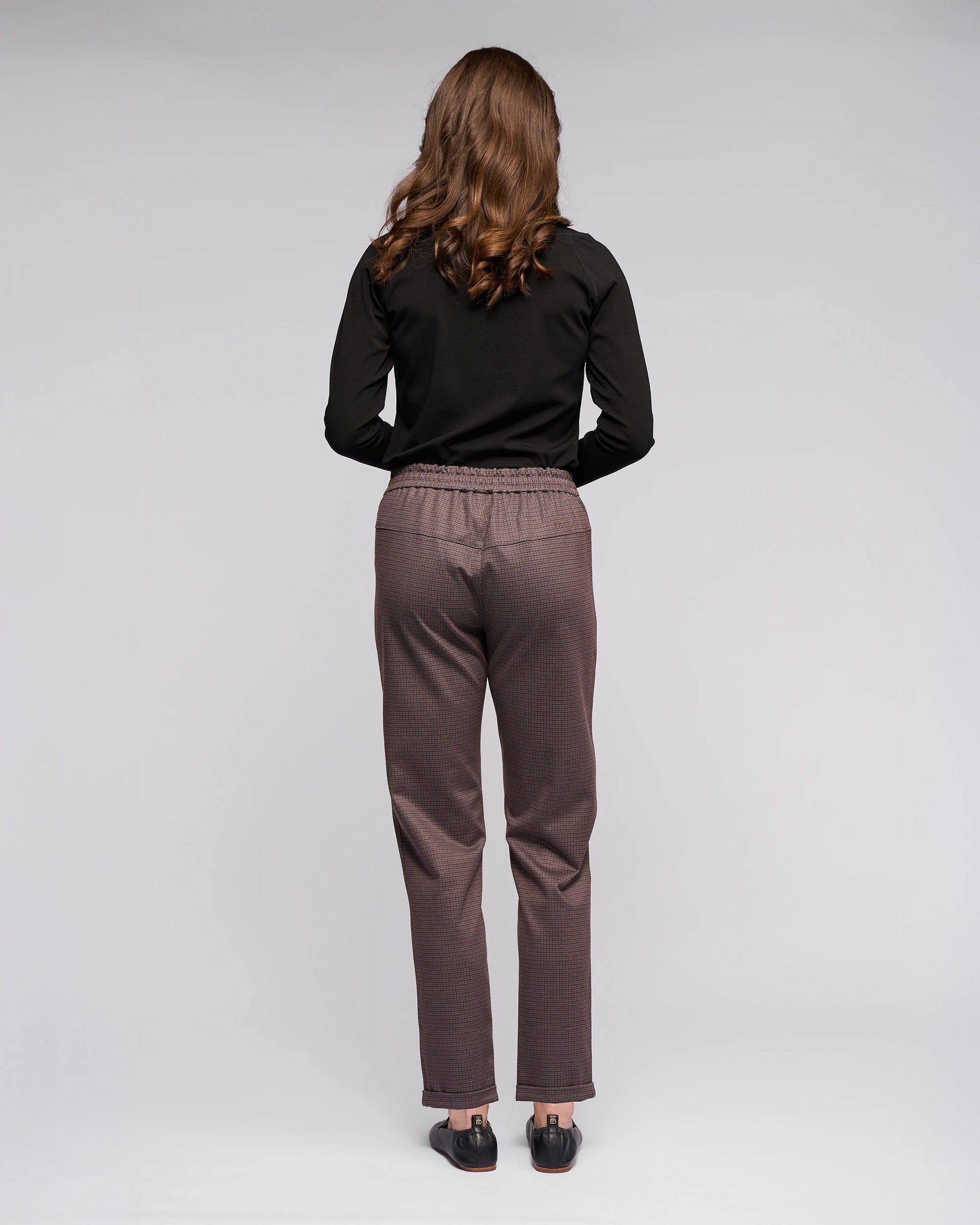 A woman with wavy brown hair, dressed in a black long-sleeve top and Vassalli Brooklyn Pant in Chocolate, stands facing away from the camera against a plain light background.