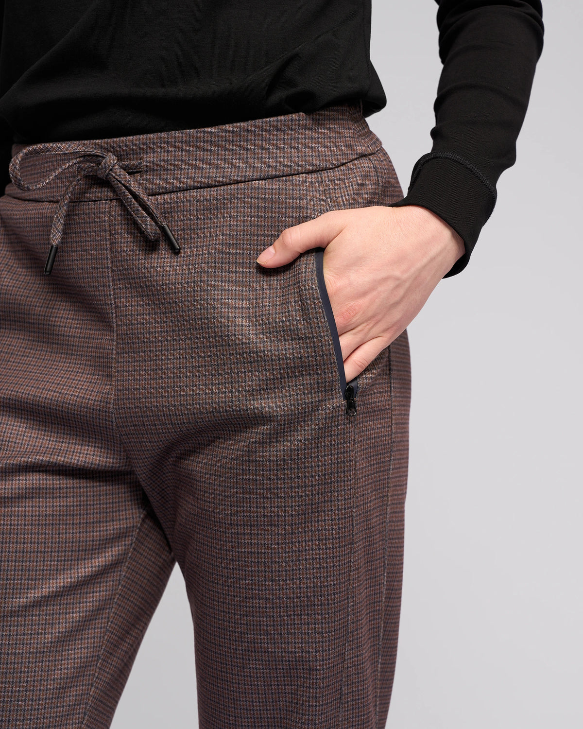 Wearing Vassalli’s Brooklyn Pant - Chocolate, a person stands with their right hand in the front zippered pocket. The relaxed-leg, ankle-grazer pants contrast against a plain, light background, paired with a black top.