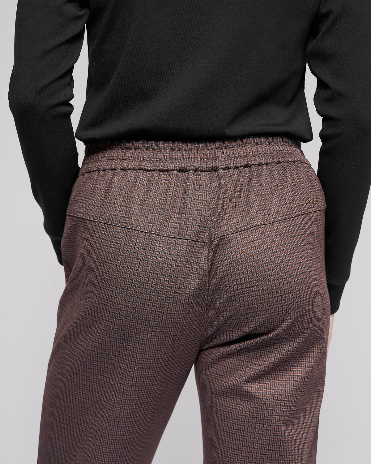 Someone is shown from the back wearing a black long-sleeve shirt and Vassalli's Brooklyn Pant in Chocolate, featuring a high-waisted, check pattern and an elastic waistband.