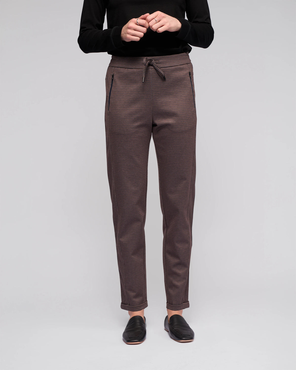 A person stands against a plain background wearing a black long-sleeve top, Vassalli's Brooklyn Pant in chocolate with zippered pockets and a drawstring waist, and black slip-on shoes. The person's head is not visible.