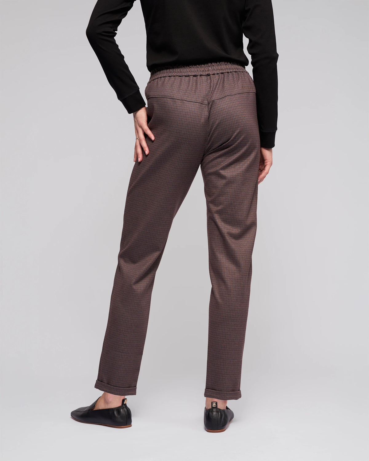 Someone stands against a plain light background, facing away with one hand on their hip, wearing Vassalli's Brooklyn Pant in Chocolate, paired with a black long-sleeve top and black flat shoes.