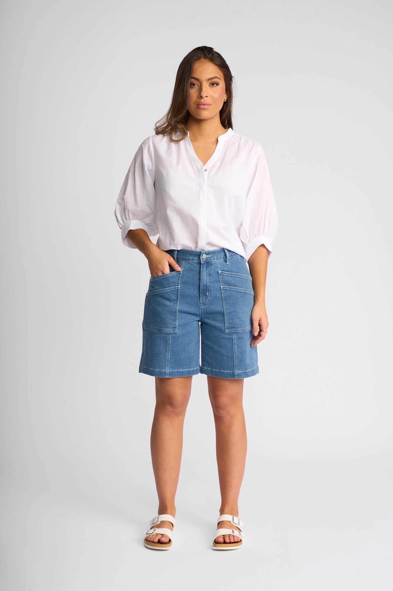 A woman stands against a plain background wearing a white blouse with puffy sleeves, the Vassalli Raven Short in Blue Hue Denim featuring large pockets and a stretch fit, and white sandals. She has one hand in her pocket and looks toward the camera.