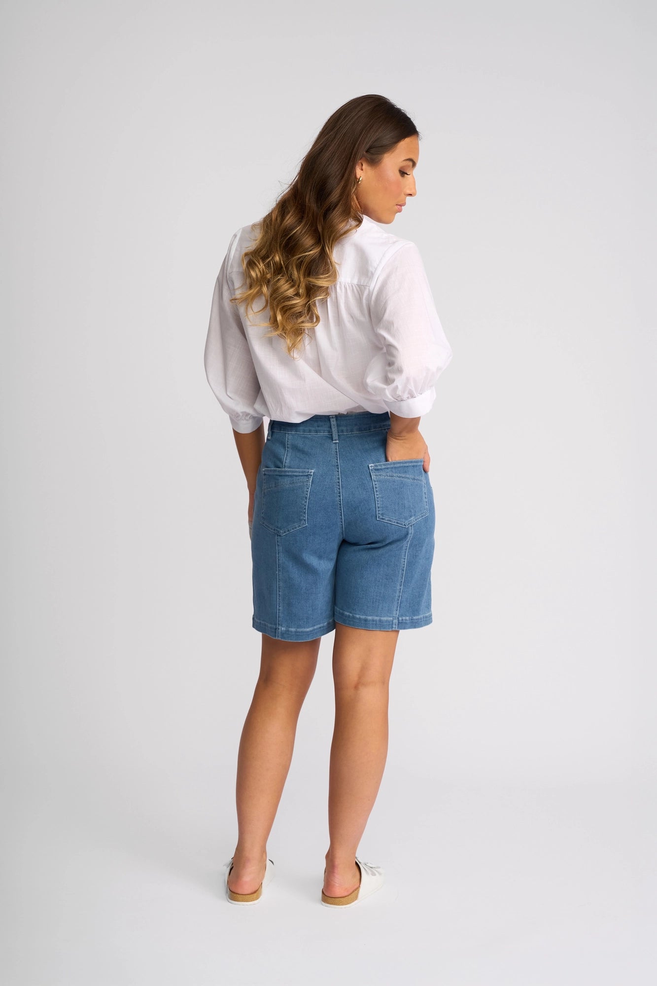 A woman with long, wavy hair stands facing away with her hands in the pockets of Vassalli’s Raven Short - Blue Hue Denim, styled with a white blouse, against a plain light background.