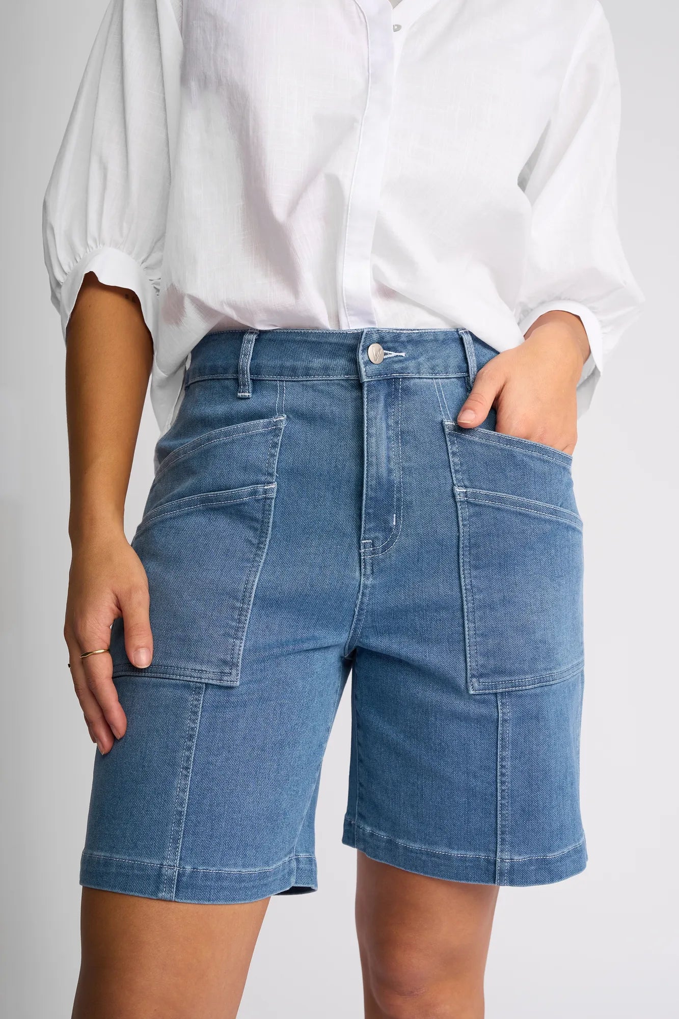 A person wearing the Vassalli Raven Short in Blue Hue Denim, paired with a white button-up shirt, stands with one hand in a pocket against a plain background. The shorts feature large front pockets and stretch denim fabric.