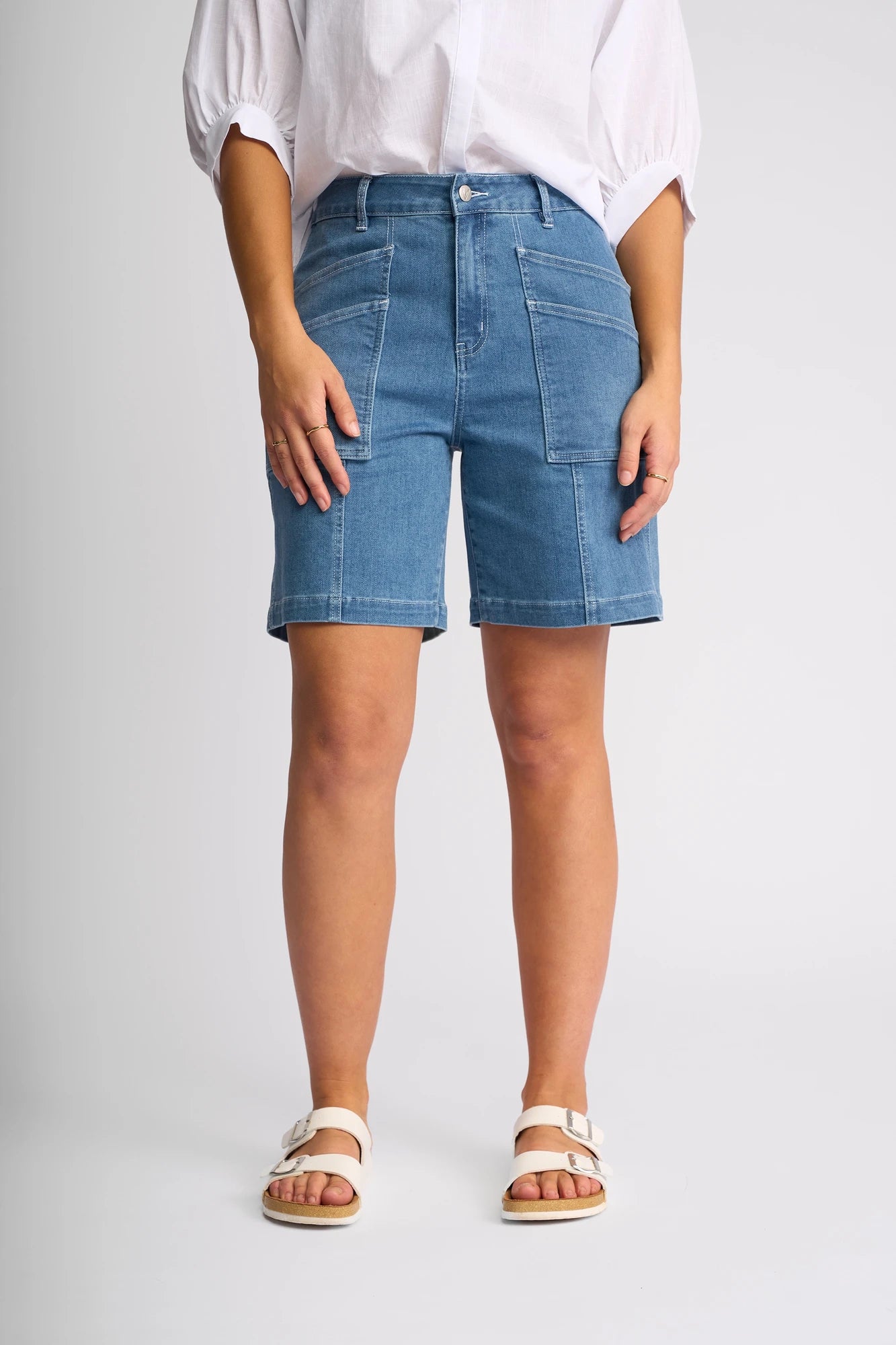 A person is shown from the waist down wearing the Vassalli Raven Short in Blue Hue Denim, styled with a white shirt and white sandals, standing against a plain light background. The stretch denim shorts feature front pockets and a classic blue hue.