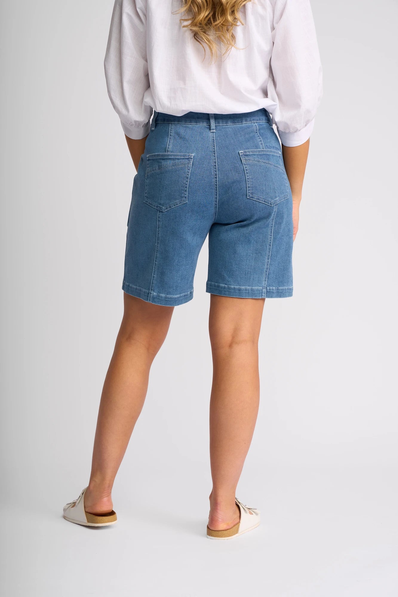 A woman stands facing away, wearing the Vassalli Raven Short - Blue Hue Denim, styled with a white blouse and beige slip-on shoes. Her wavy blonde hair cascades over her shoulders, set against a plain, light-colored background.