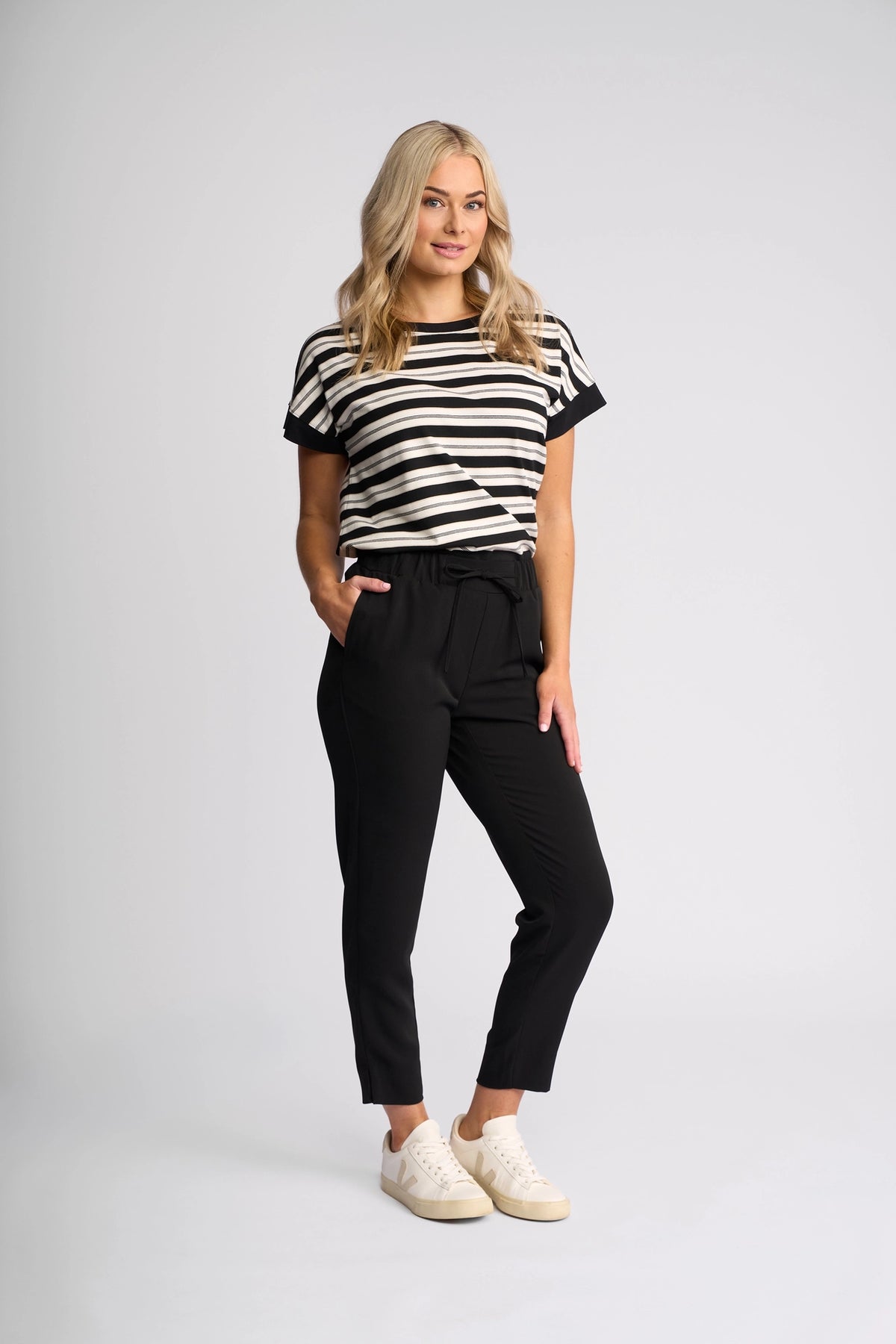 A woman with long blonde hair stands smiling, dressed in a black and white striped t-shirt and the Vassalli Marble Pant - Black for lightweight comfort and effortless style, paired with white sneakers against a plain light background—making these Vassalli pants the perfect travel companion for any adventure.