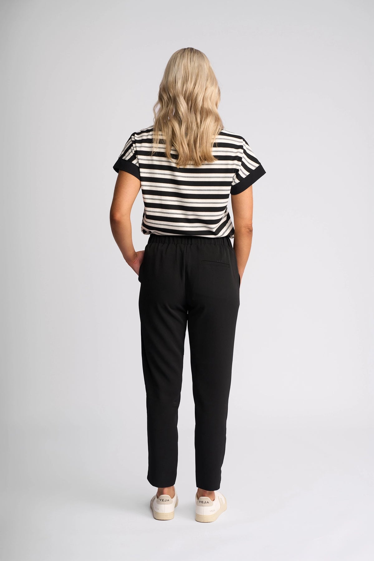 A person with long blonde hair stands facing away, wearing a black and white striped shirt, the Vassalli Marble Pant in Black for lightweight comfort, and white sneakers against a plain light background—a perfect look for any travel companion.