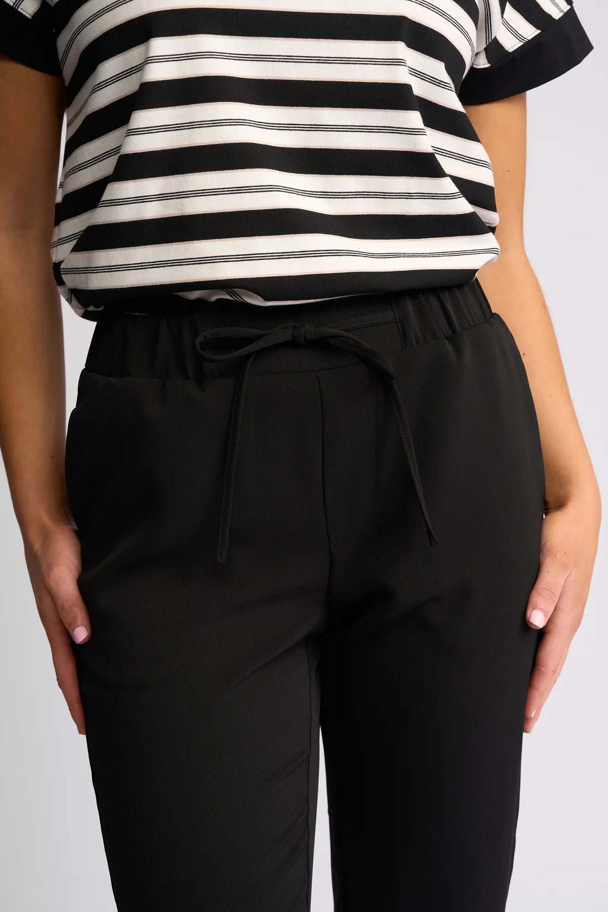 A person wearing a black and white striped short-sleeve shirt and black drawstring pants stands with arms relaxed by their sides. The image, cropped from shoulders to mid-thigh, showcases the lightweight comfort of the Vassalli Marble Pant - Black—a perfect choice for effortless travel style.
