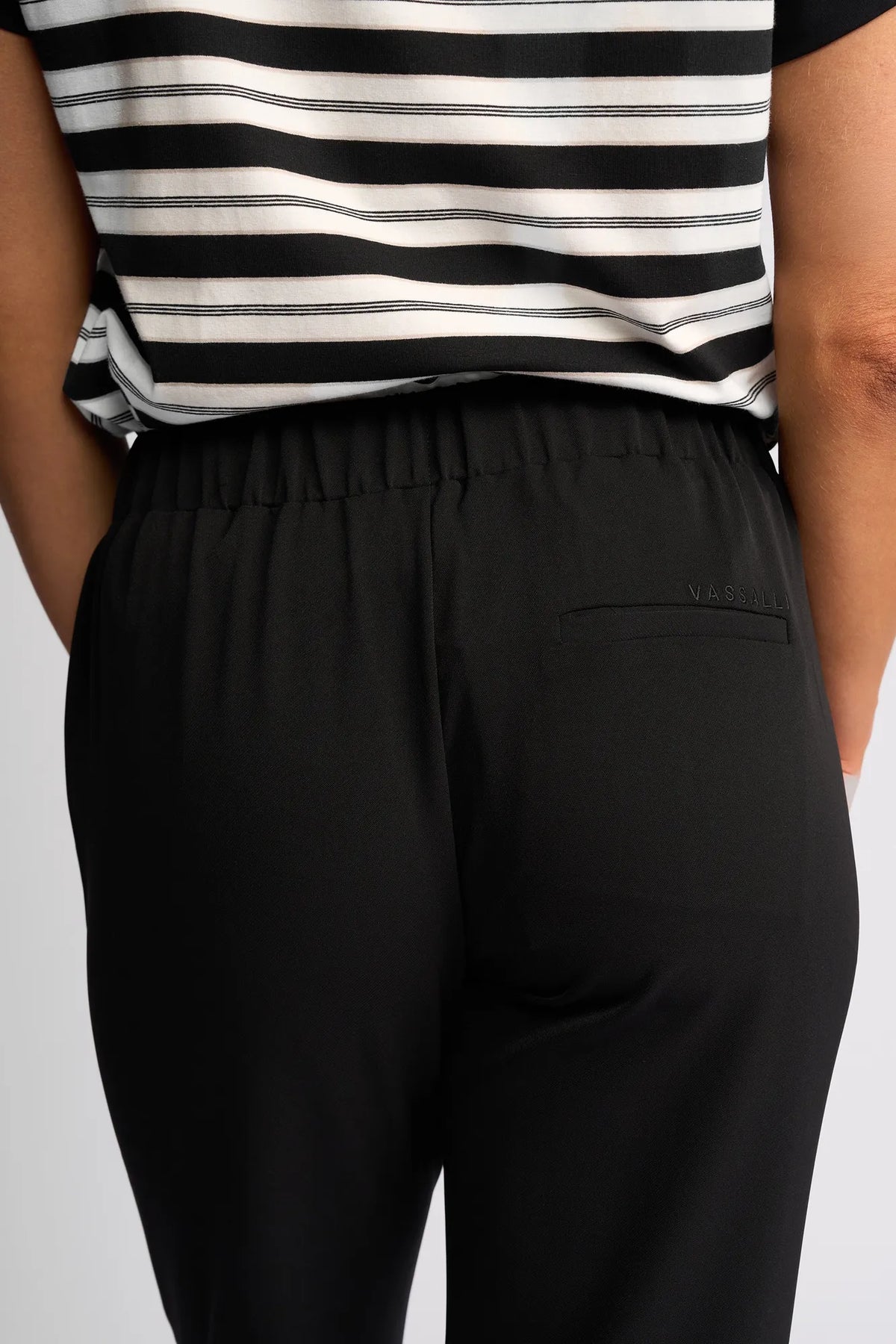 A person with their back to the camera, shown from the waist down, wears Vassalli’s Marble Pant - Black. This style features a single back pocket and an elastic waistband for lightweight comfort—making it an ideal travel companion.