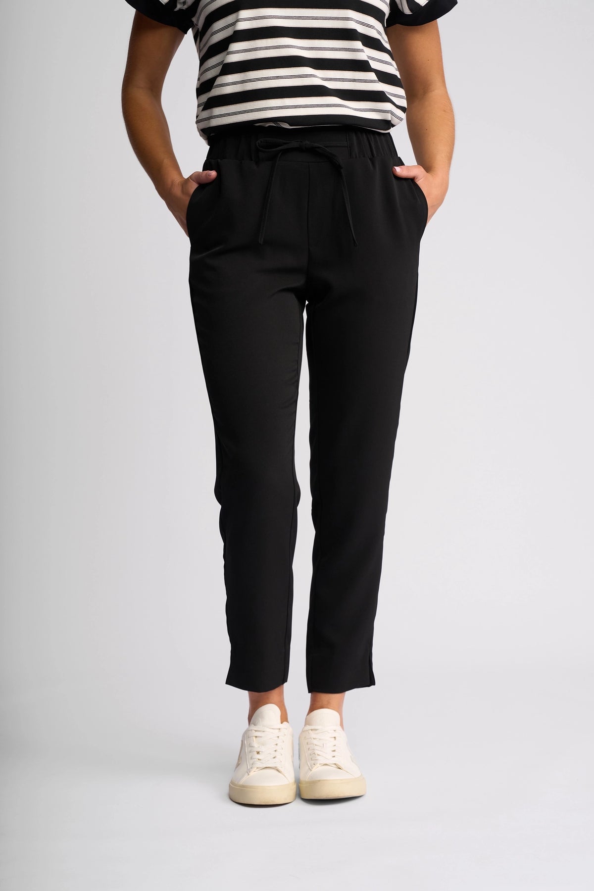 A person wearing a black and white striped shirt, the Vassalli Marble Pant in Black—renowned for its lightweight comfort—and white sneakers, stands with hands in pockets against a plain light gray background.