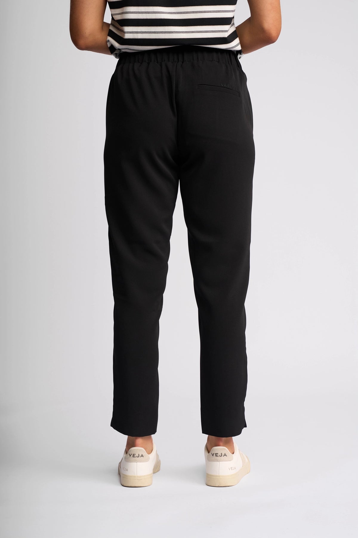 A person stands facing away, wearing the Vassalli Marble Pant in black, paired with a black and white striped top and white Veja sneakers, against a plain light background.